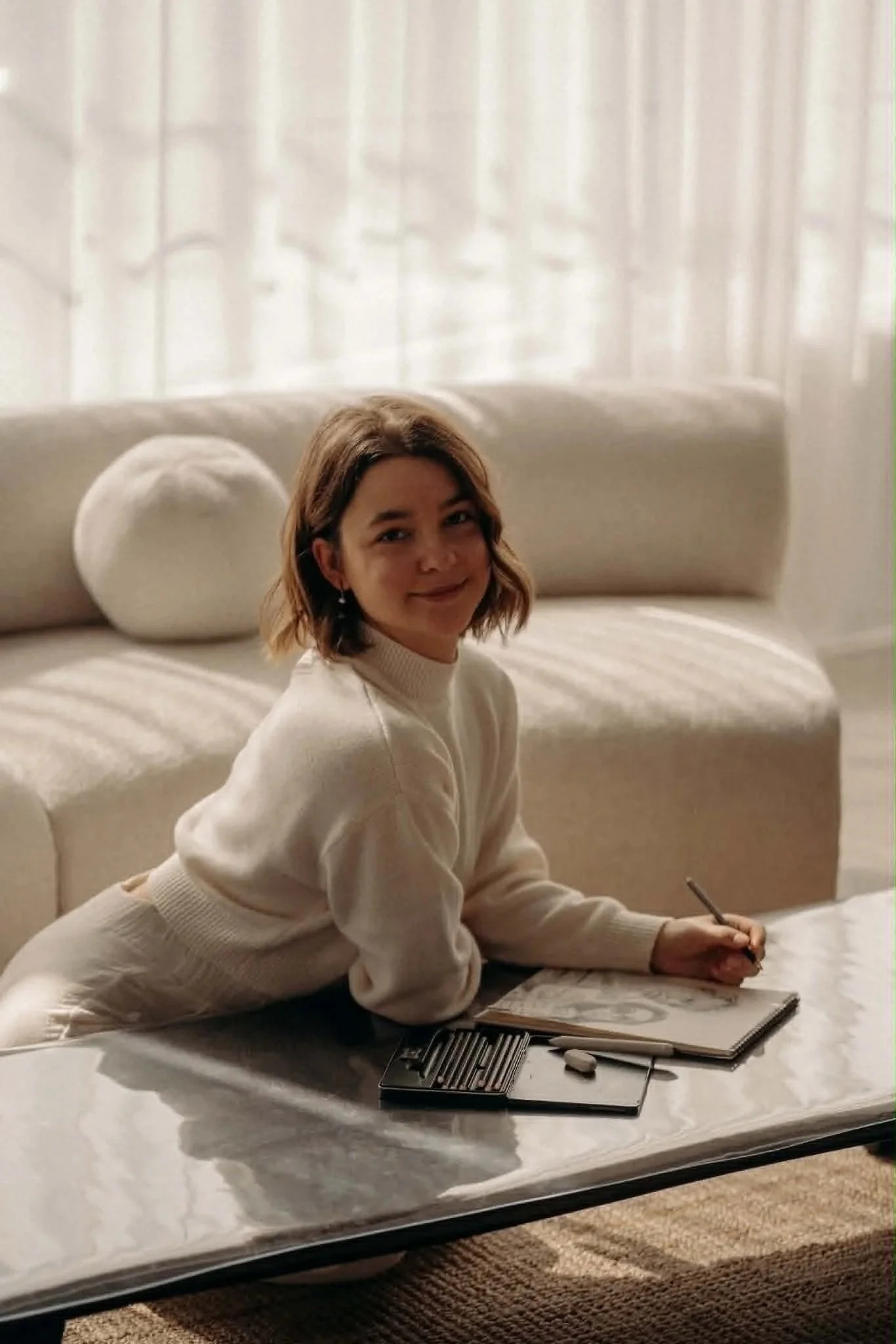 Une jeune femme assise sur un lit, en train de dessiner ou écrire avec un stylo, avec un maquillage et un sourire discret, dans une chambre avec des rideaux blancs en arrière-plan.