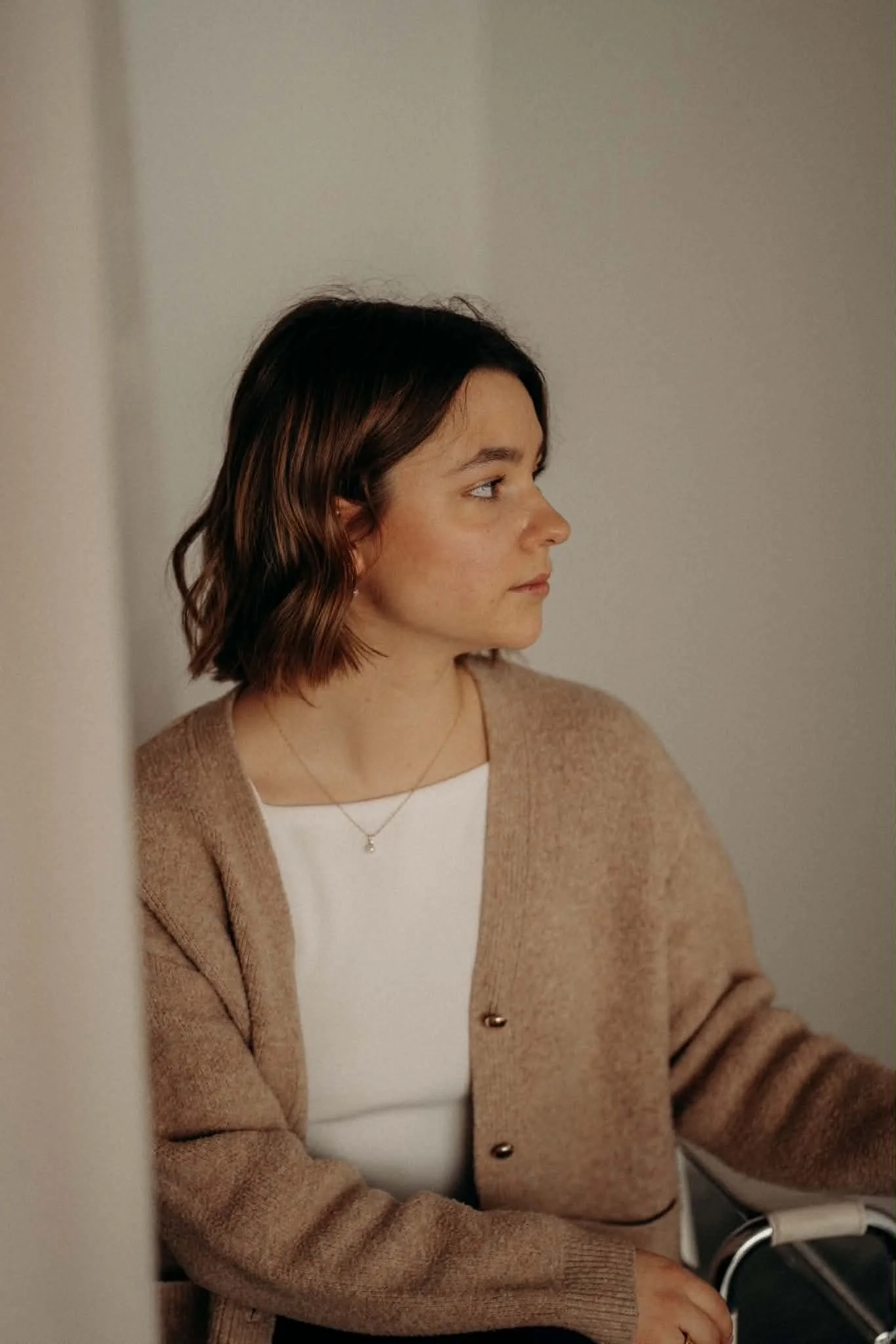 Portrait d'une femme aux cheveux bruns, coiffée en vagues, portant un chandail beige et un collier suspendu. Elle regarde vers la droite, dans un environnement intérieur.
