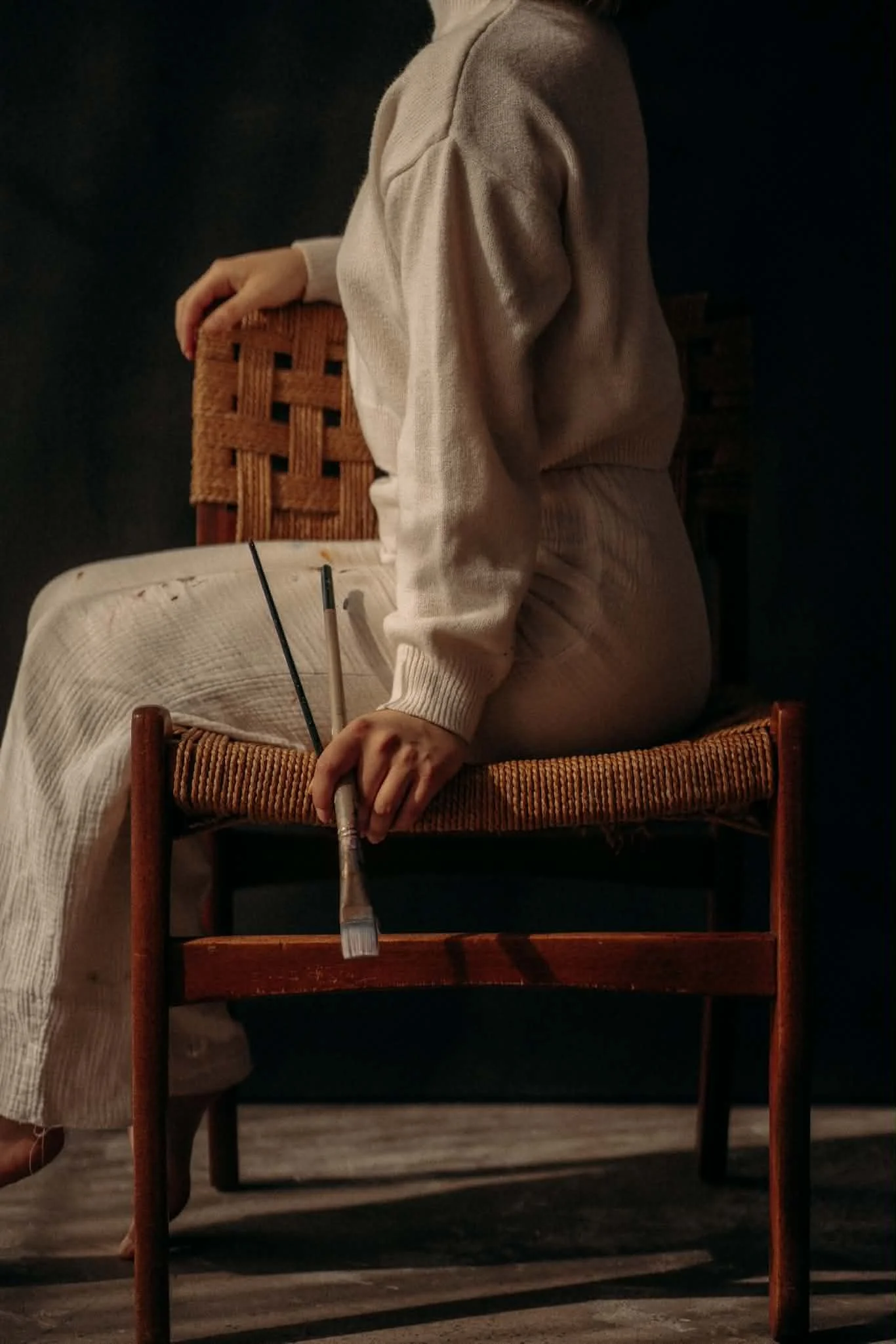 Une personne assise sur une chaise en bois, vêtue d'un pull et de pantalon kaki, tenant une baguette. Le fond est sombre, ambiance chaleureuse.