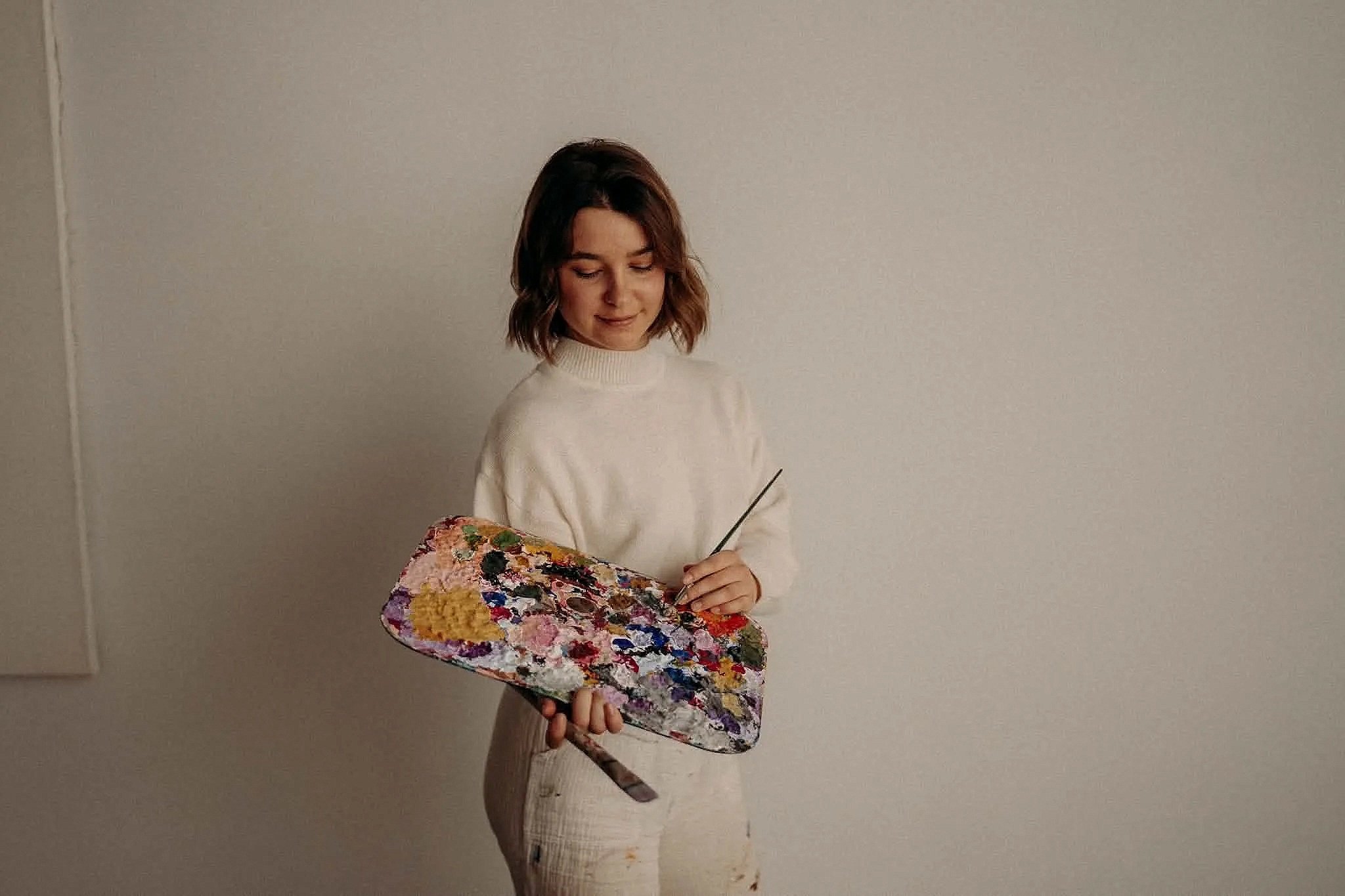 Une femme avec des cheveux bruns et courts, portant un pull blanc, tient une palette de peinture colorée et un pinceau, se tenant devant un mur beige.