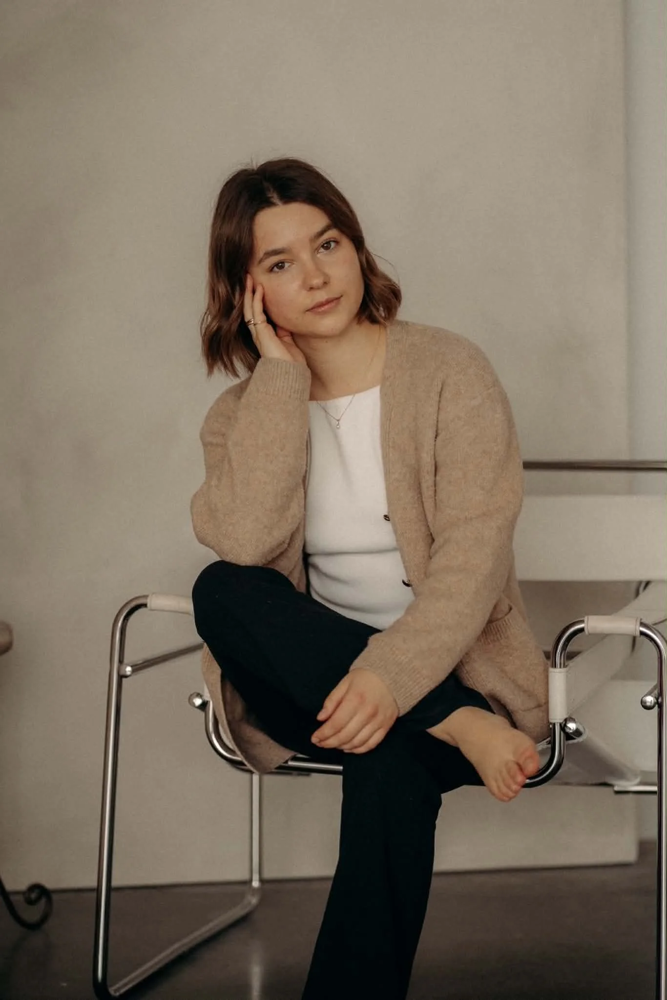Une femme assise sur une chaise, pieds nus, en équilibre, avec une expression neutre, portant un chandail beige et un t-shirt blanc, dans une pièce avec un mur clair.