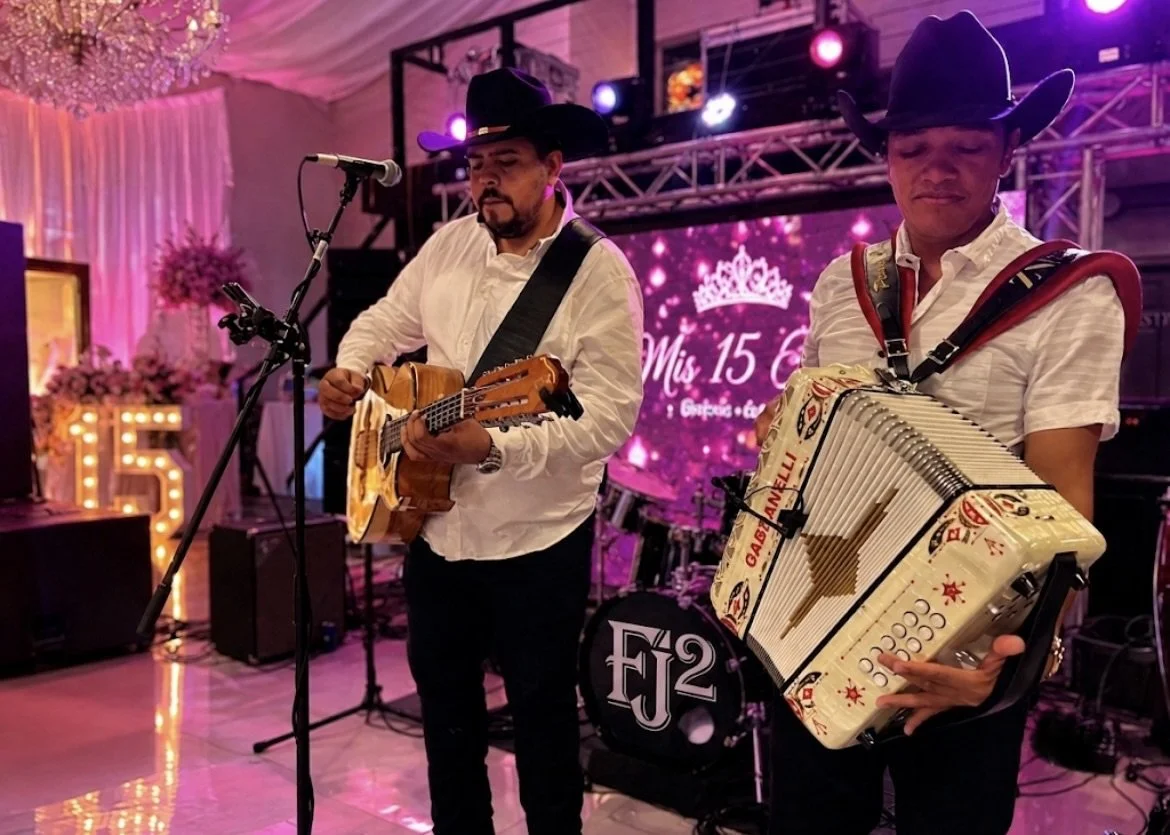 Two men wearing cowboy hats and white shirts performing with a guitar and an accordion on stage at a quinceañera celebration.