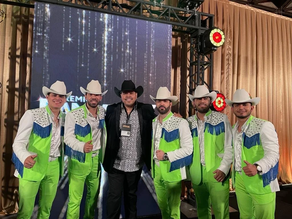 Six men dressed in matching neon green and white cowboy costumes with fringe, hats, and silver embellishments, standing on stage with one man in black attire, all smiling at the camera.