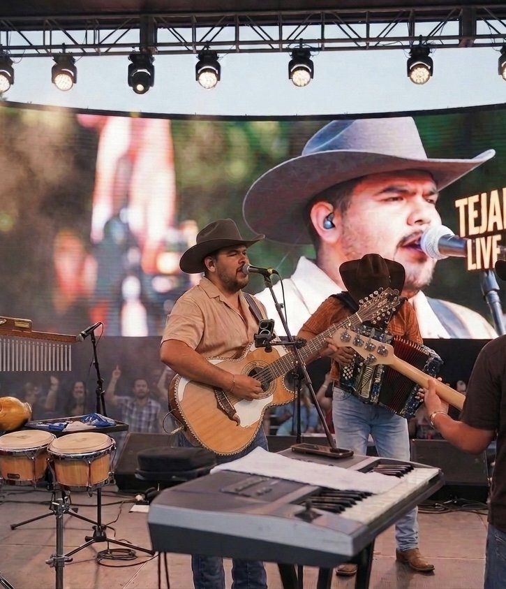 Musicians performing live on stage, wearing cowboy hats, with a large screen behind them showing a close-up of one singer singing into a microphone.