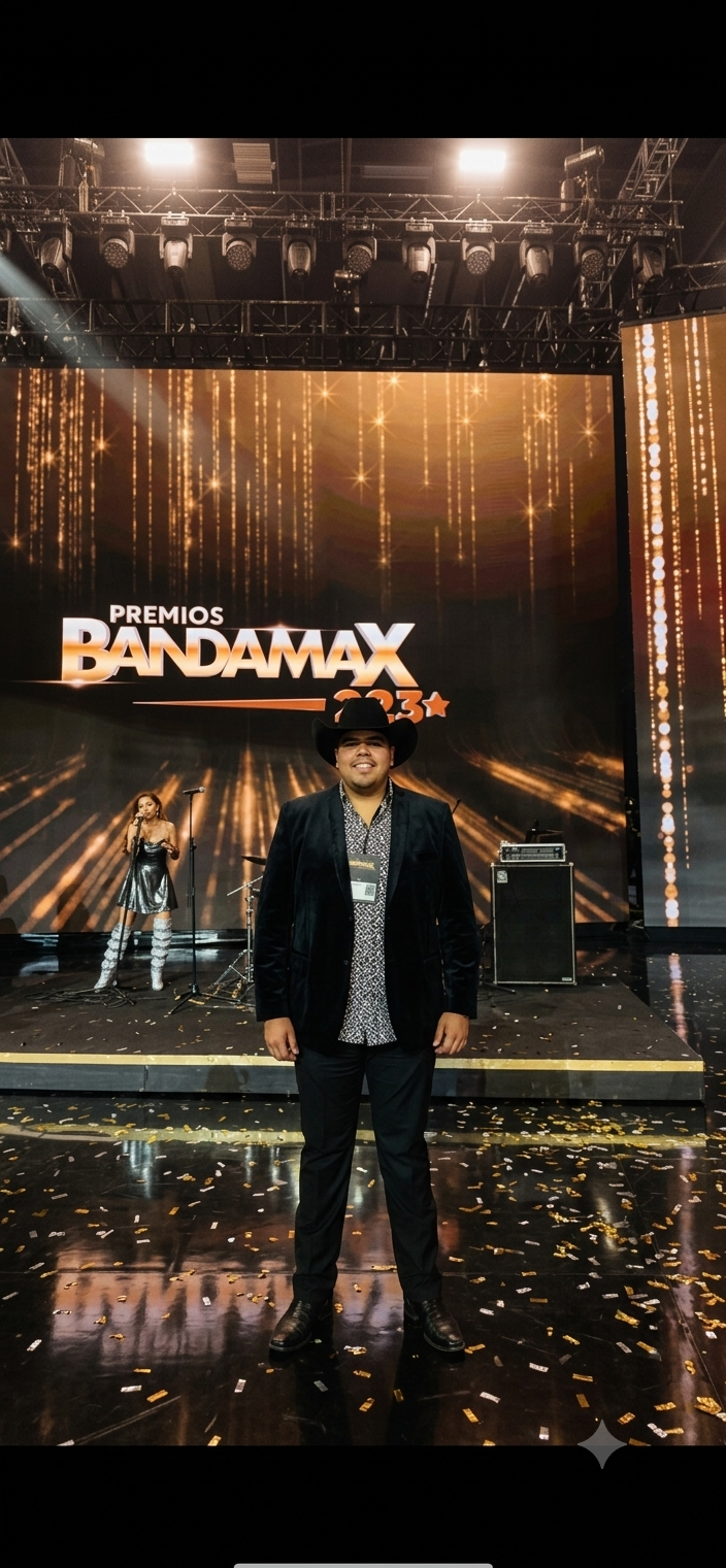 Man in a black hat and suit standing on a stage at the Premios Bandamax 2023 awards ceremony, with a background featuring a large screen displaying the event's logo and a female performer in the distance.