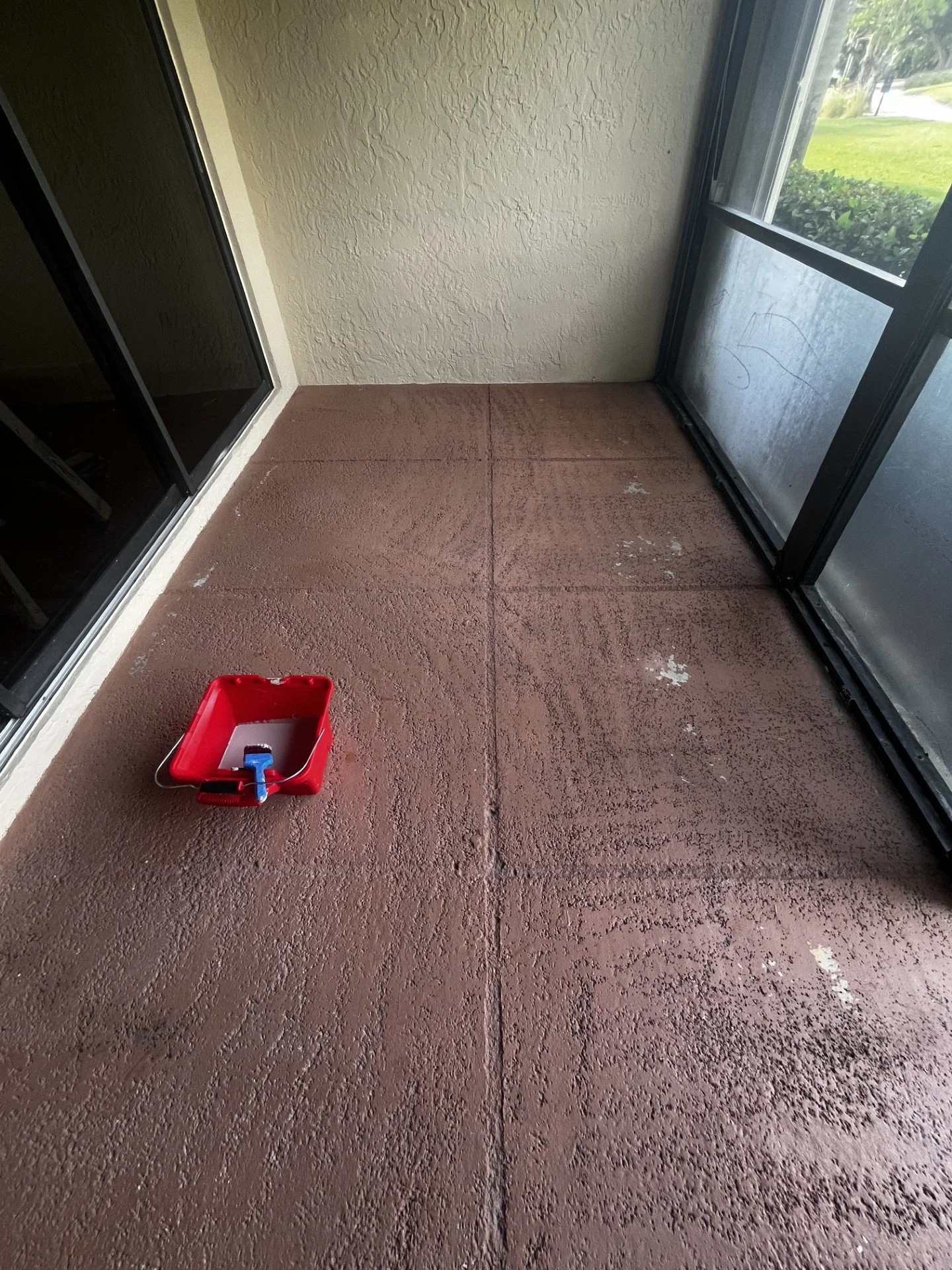 A small red paint tray with a roller inside sits on a textured reddish-brown concrete porch. The porch is enclosed by a screen door and a glass door, with greenery visible outside.