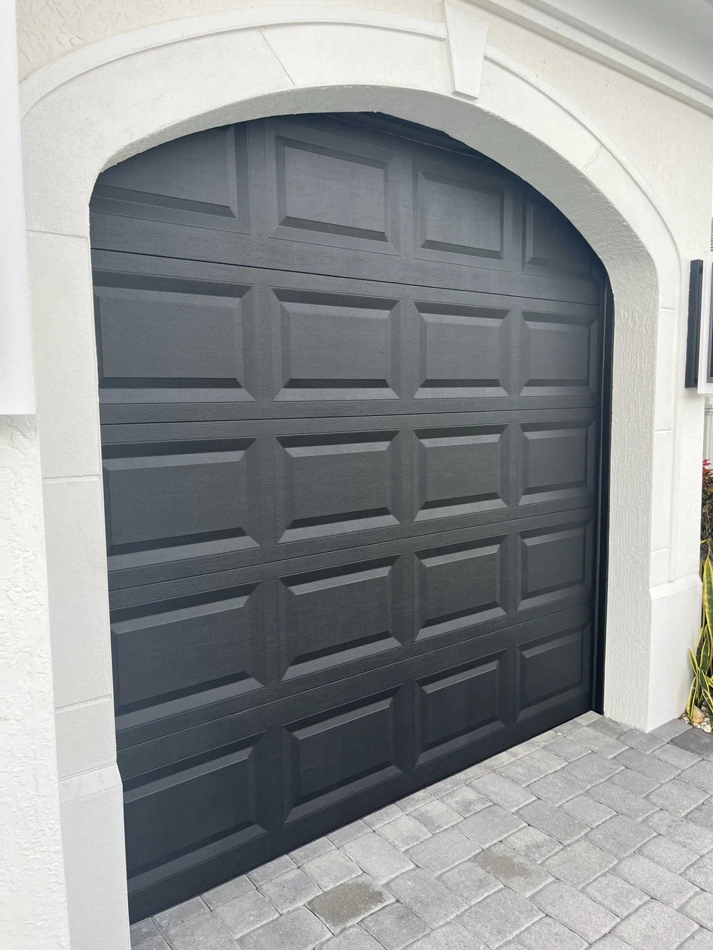 Black sectional garage door with a geometric panel design, surrounded by white stucco walls and gray stone pavers in front.