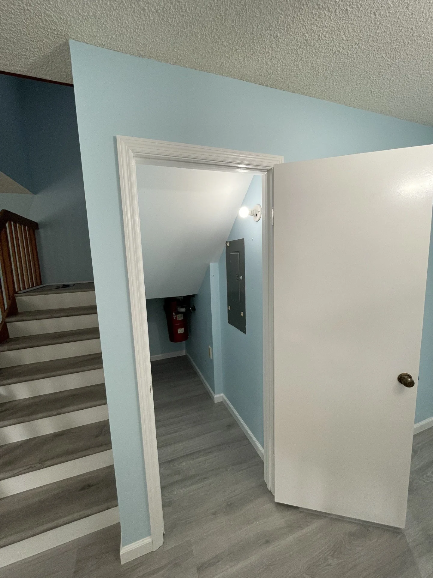 Interior view of a hallway with stairs on the left, light blue walls, white door partially open, and a red fire extinguisher mounted on the wall.
