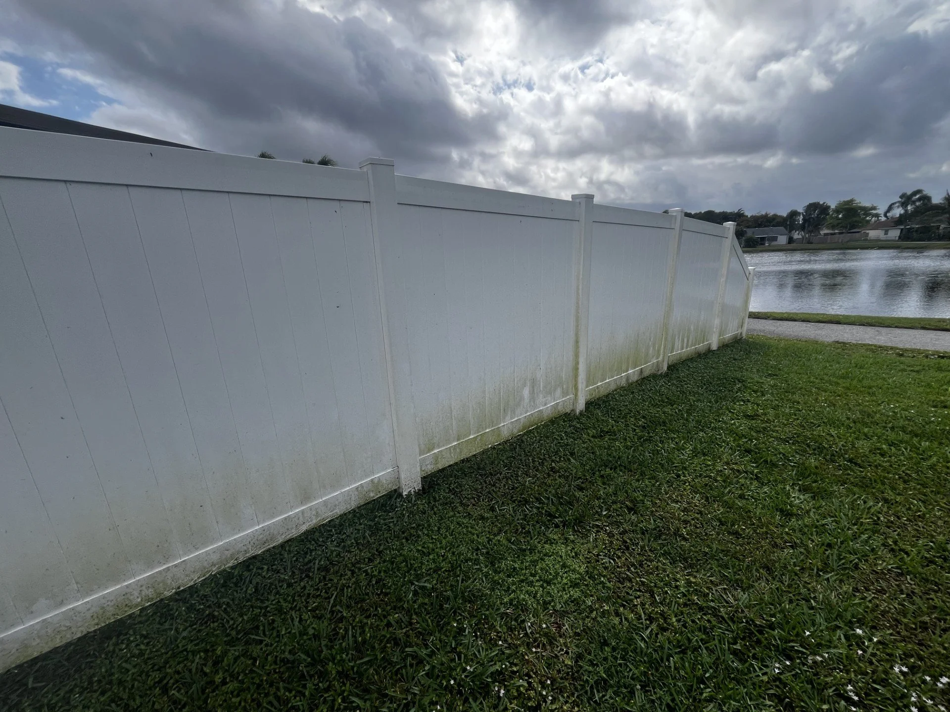 A white fence along a grassy area near a body of water with houses in the background, under a cloudy sky.