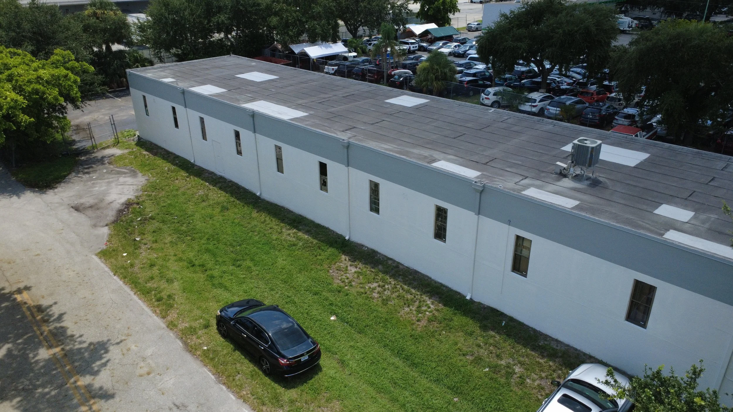 Aerial view of a white building with a flat roof, surrounded by trees and parking lot filled with cars.