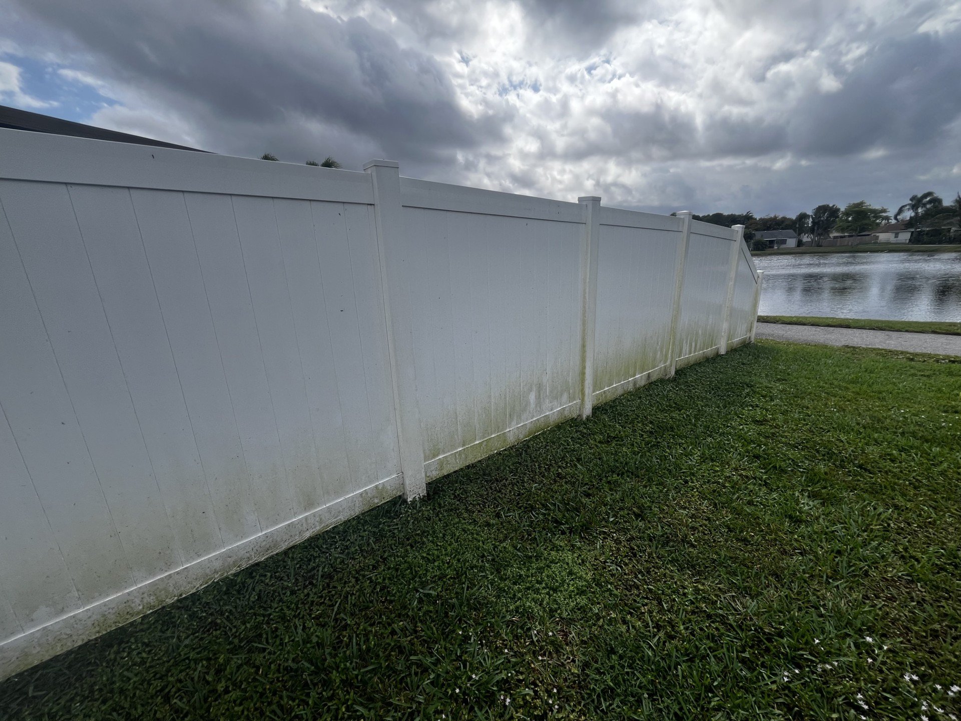 A white vinyl fence runs alongside a grassy area near a body of water, with houses and trees visible in the background under a cloudy sky.