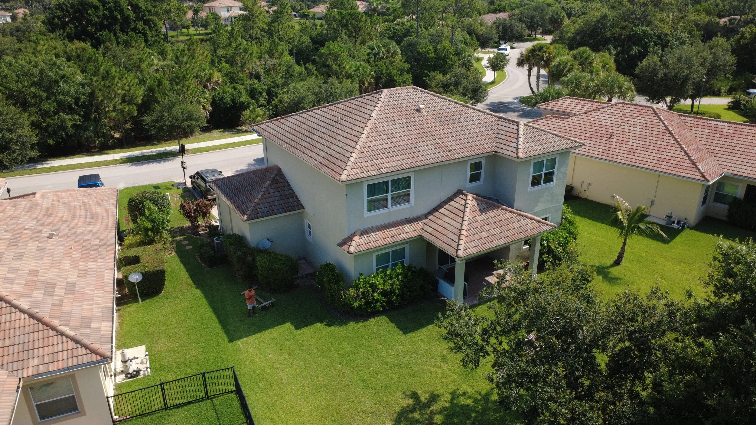 Aerial view of a two-story house with a tiled roof, surrounded by a green lawn, trees, and neighboring houses in a suburban neighborhood.
