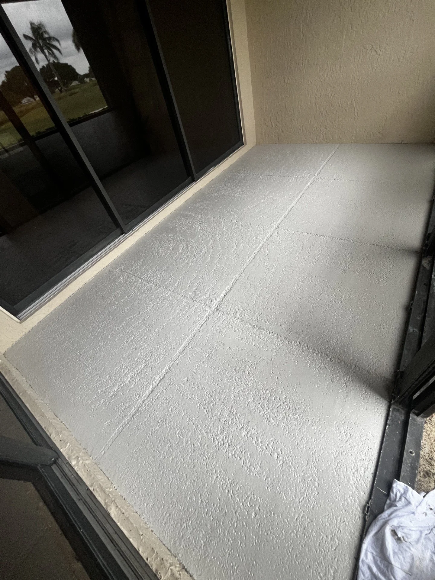 Photo of a balcony with newly painted white textured concrete floor, sliding glass door, beige stucco walls, and a white cloth or paper on the floor near the door.