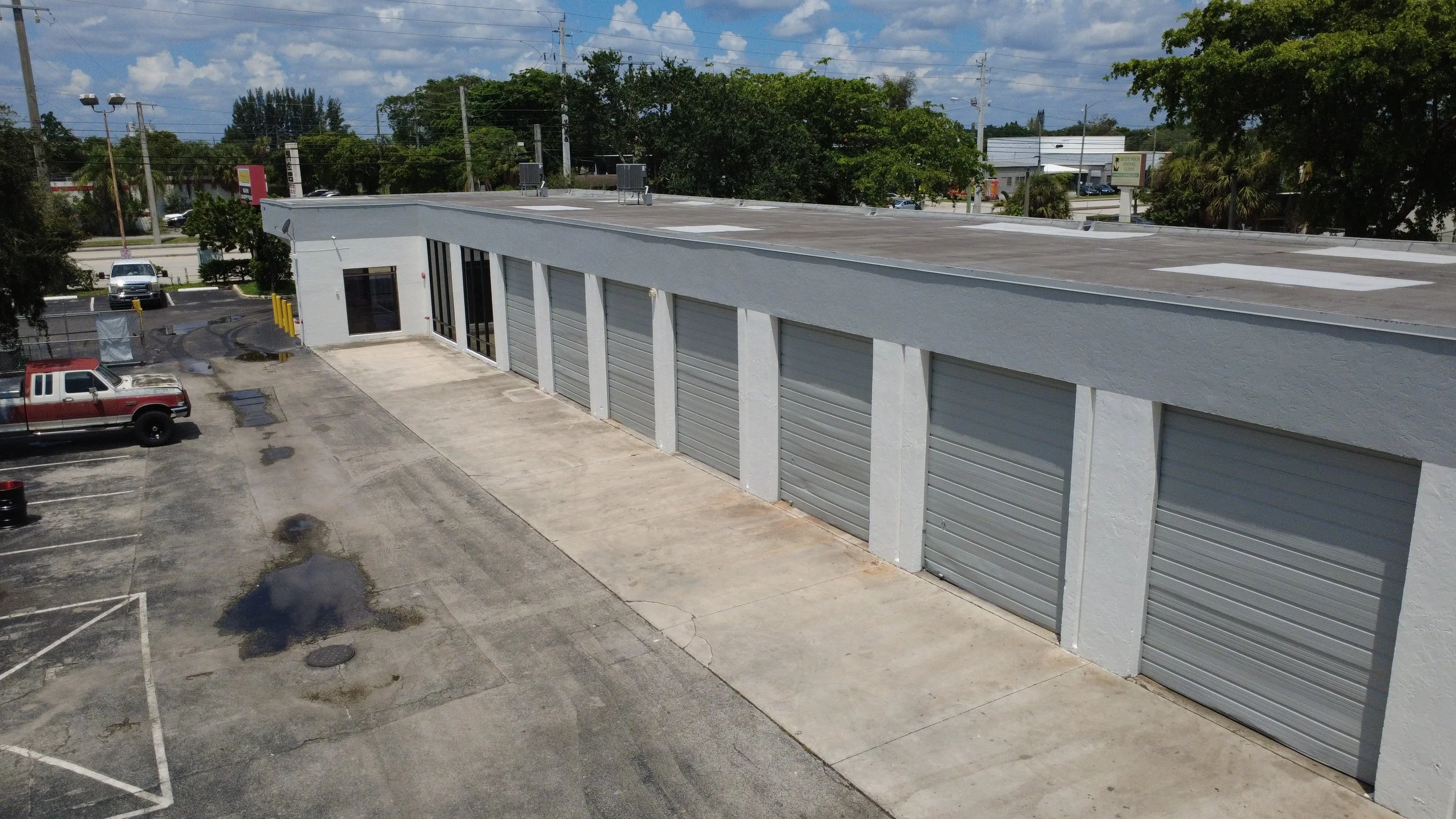 A large commercial building with multiple gray metal garage doors and a flat roof, surrounded by parking spaces and some vehicles, with trees and power lines in the background under a partly cloudy sky.