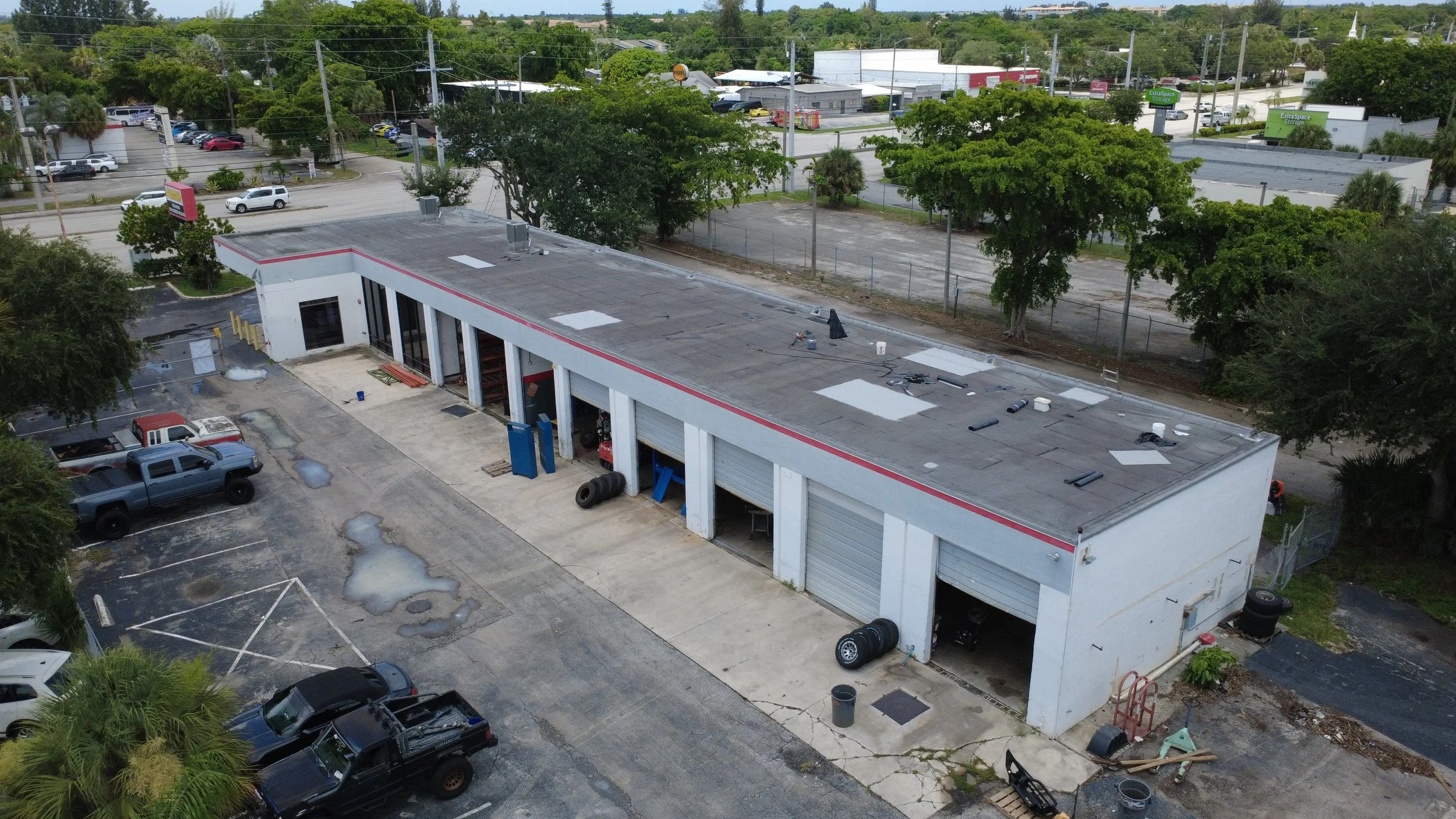 Aerial view of a commercial building under renovation, with tools and materials on the roof, surrounded by parking lots with several cars, trees, and nearby commercial establishments.