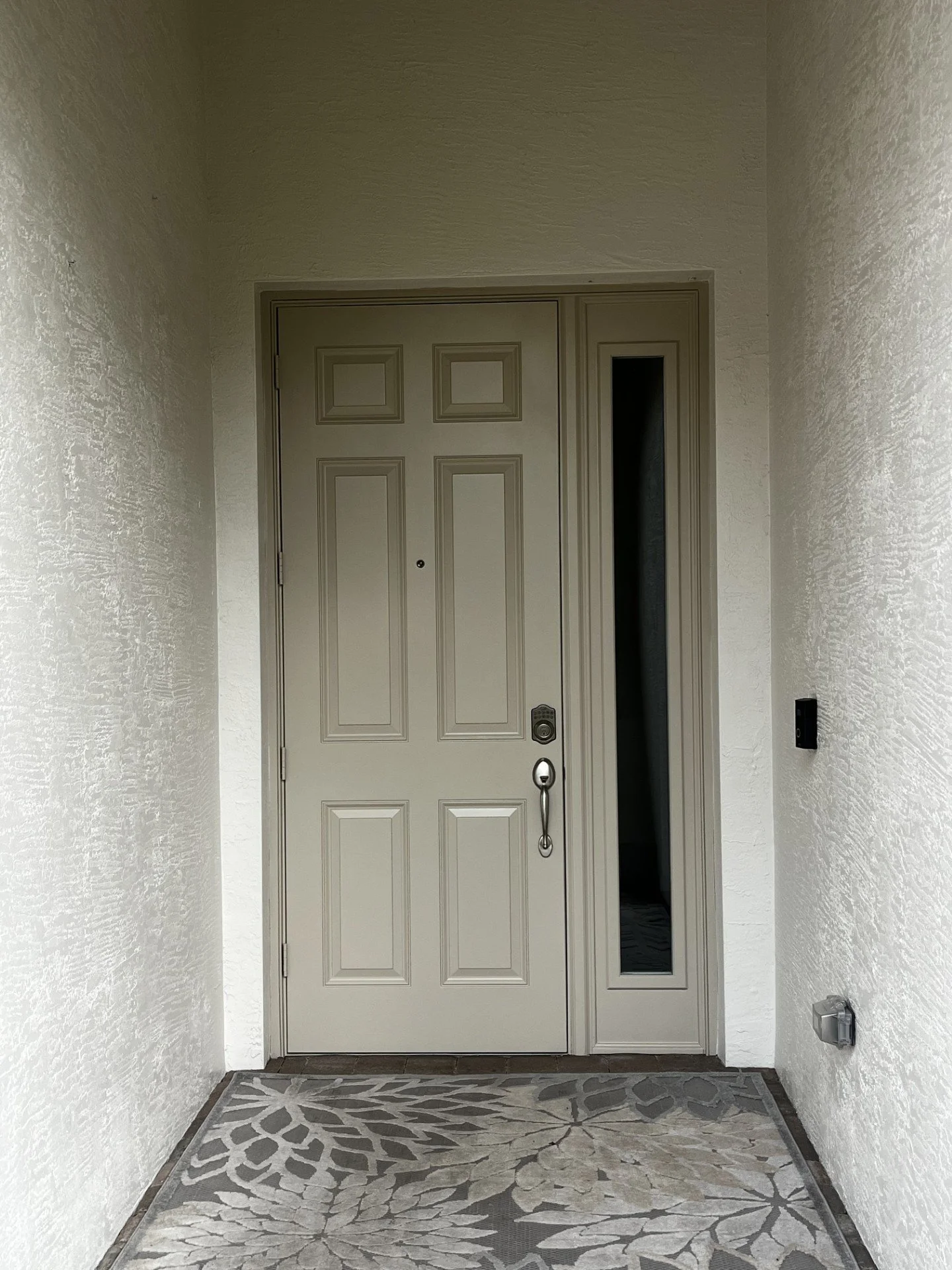 Front door with locking handle, and a small vertical window to the right, inside an entryway with textured white walls and patterned floor mat.