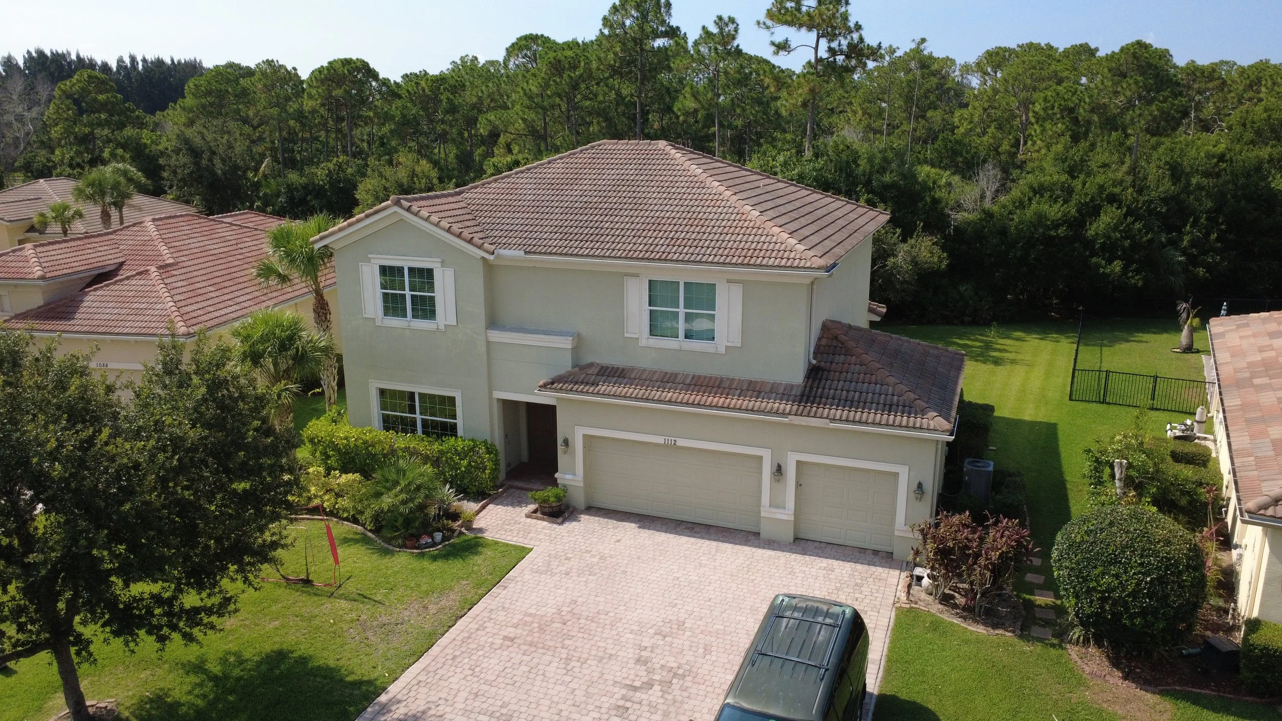 A two-story house with a beige exterior, brown tiled roof, and attached garage on a paved driveway. The front yard has green grass, small trees, and shrubs. The backyard is fenced with a well-maintained lawn and some trees, with a dense forest area v