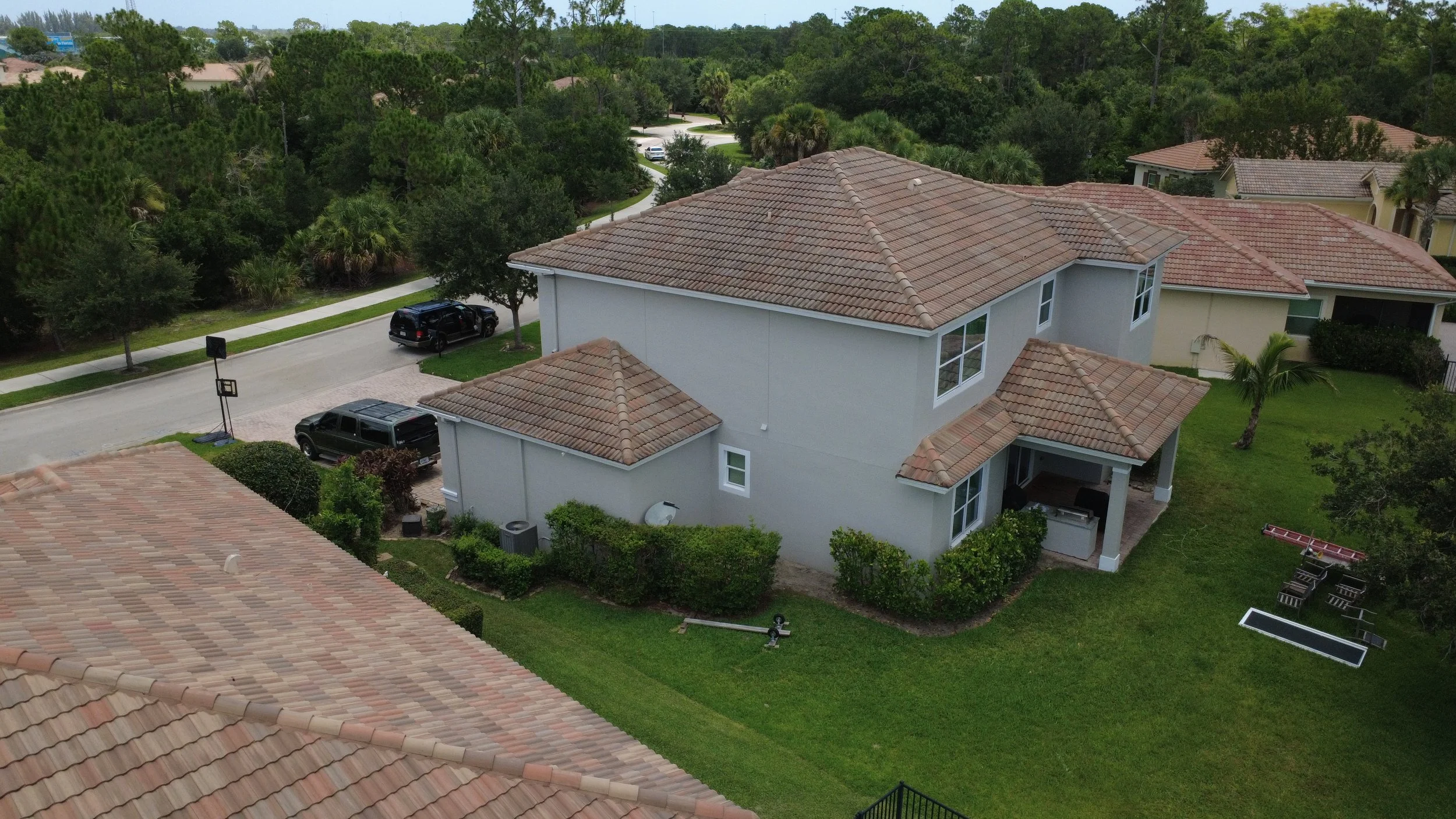 Aerial view of suburban neighborhood featuring a white house with a terracotta tile roof, surrounded by green lawns, trees, and parked cars on the street.