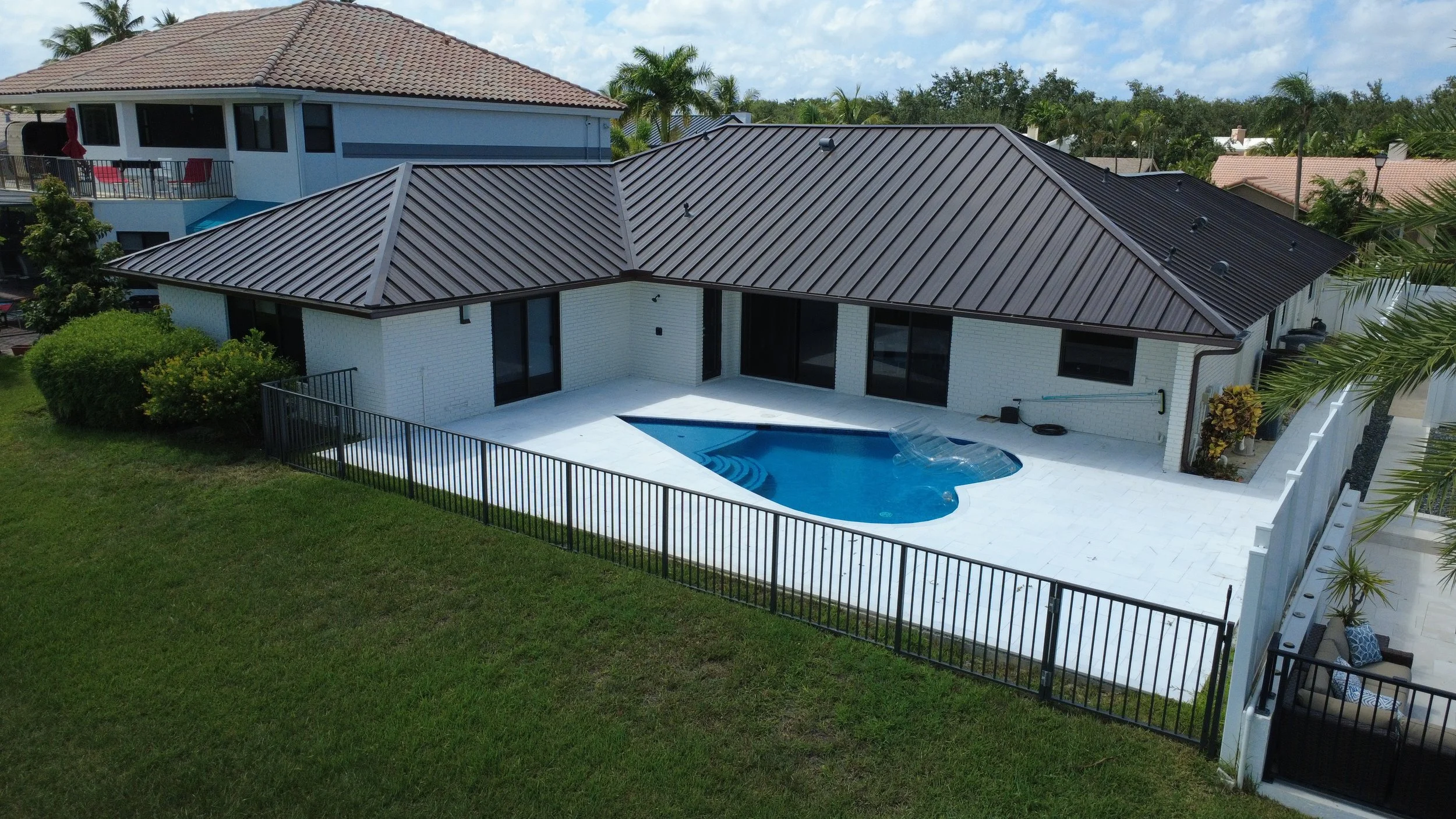 A backyard with a small swimming pool surrounded by white patio tiles, enclosed by a black metal fence. The house has white brick walls, large glass sliding doors, and a dark-colored metal roof. There are some outdoor plants and bushes around the yar