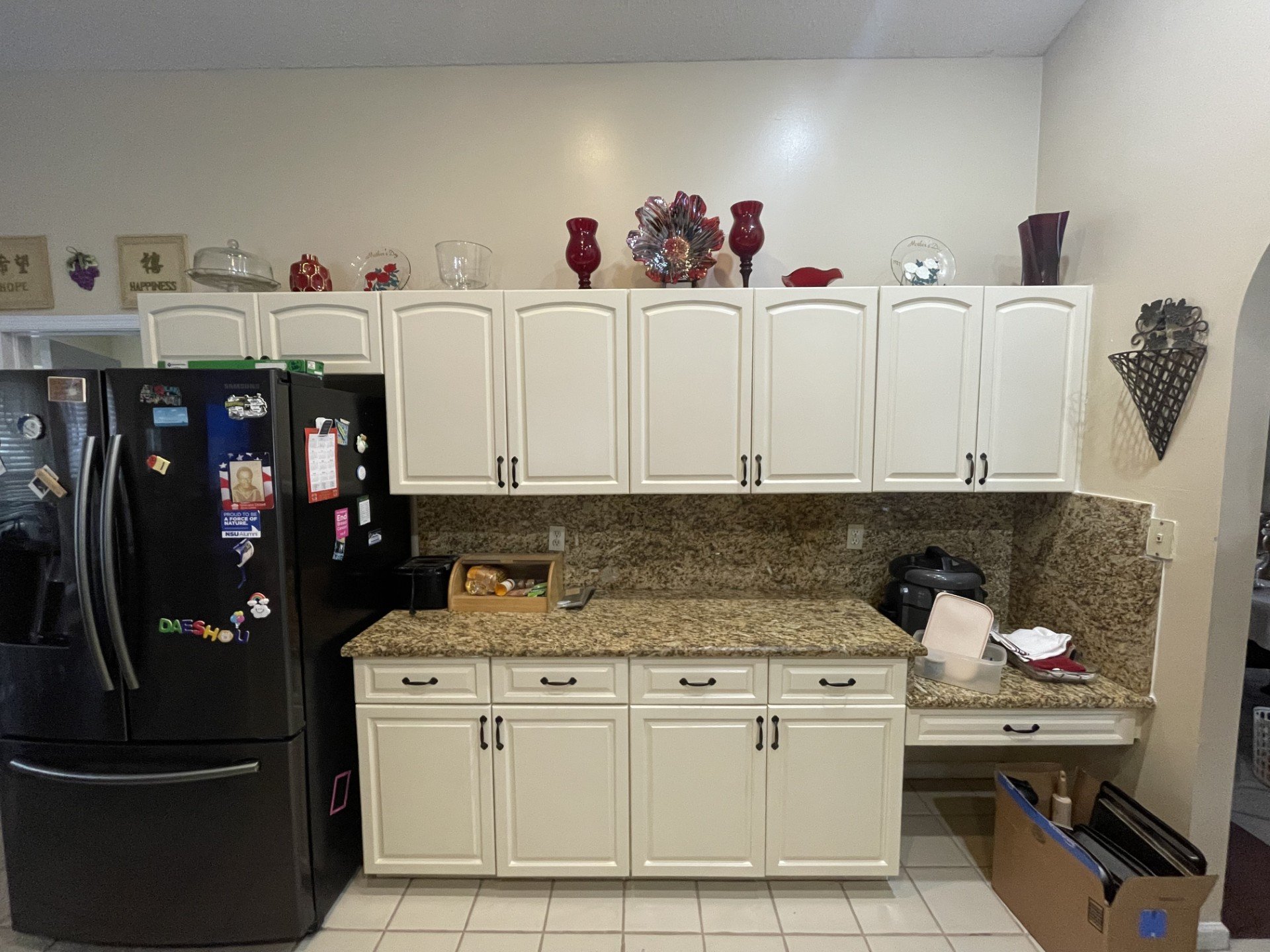 Kitchen with white cabinets, granite countertops, and various decorative items on top of the cabinets, including vases and glassware. There is a black refrigerator with magnets and stickers, and a countertop with a small wooden organizer, a plastic c