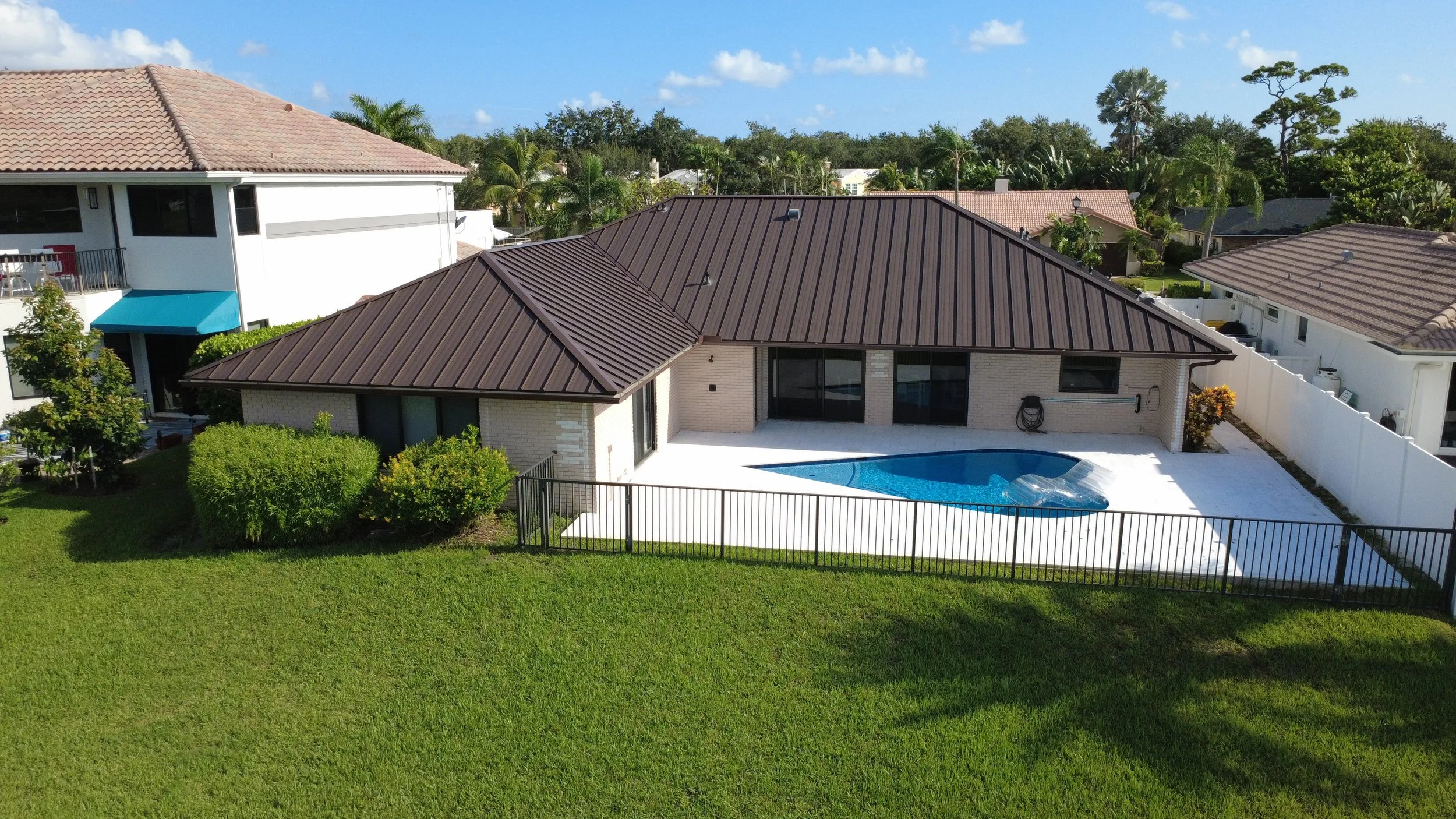 Rear view of a modern house with a backyard pool, surrounded by a white fence, with neighboring houses and lush green trees in the background under a partly cloudy sky.