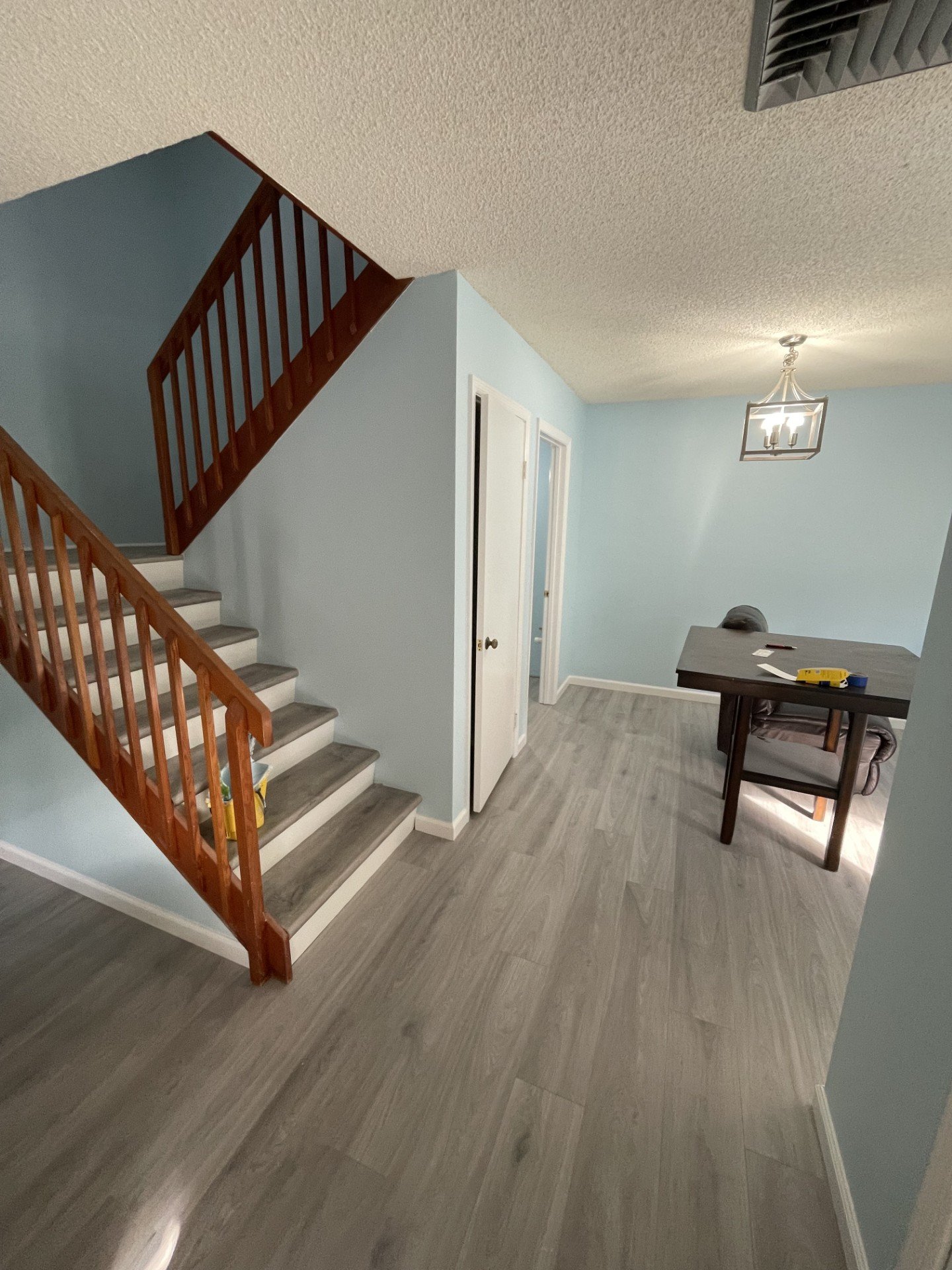 Interior view of a living area showing staircase with wooden railing, light blue walls, a white door, a table with items on it, a chair, and a pendant chandelier.