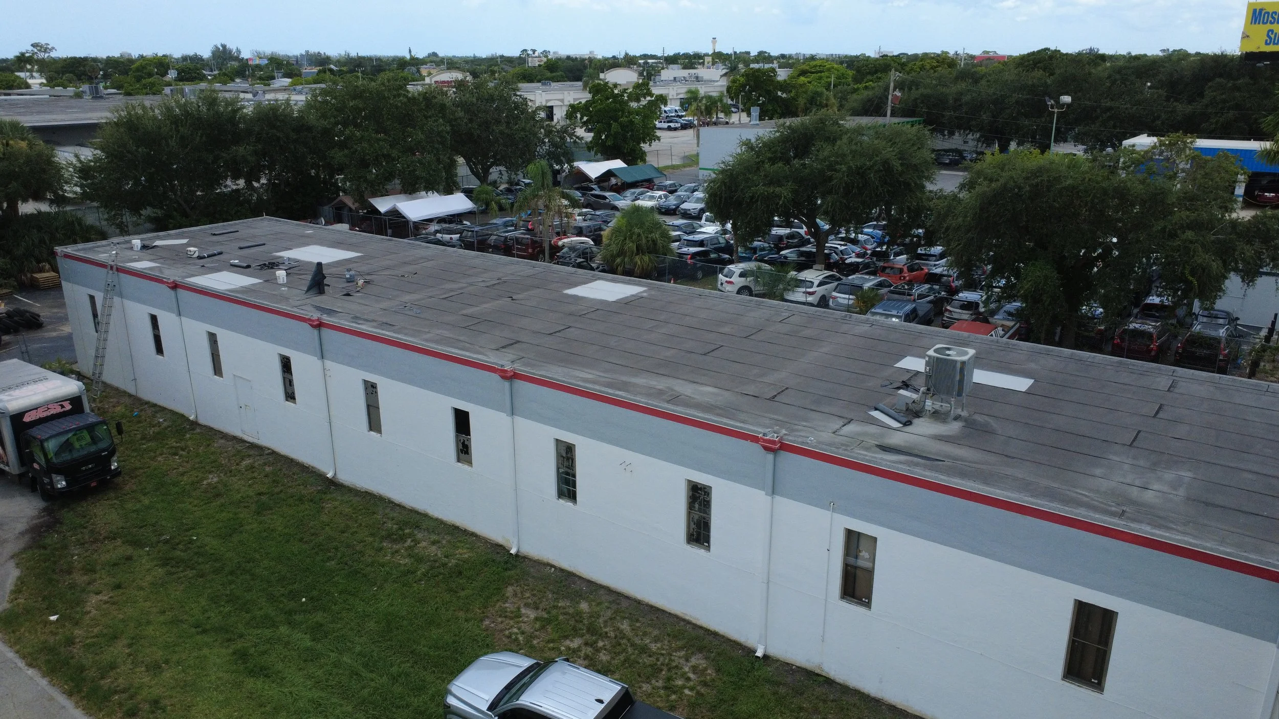 Aerial view of a flat commercial building with a parking lot and several cars, surrounded by trees and other commercial buildings.