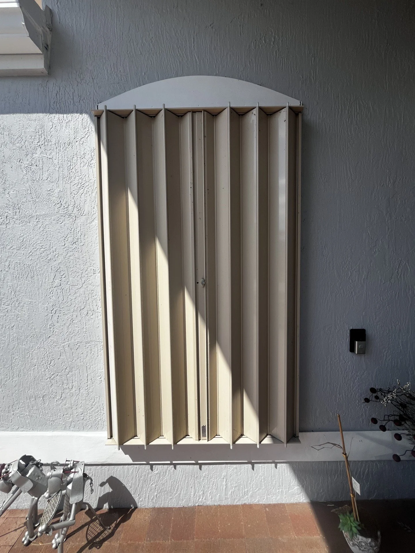 A beige exterior security shutter mounted on a white wall outdoors, with a small window with blinds above and a black light switch nearby. Sunlight and shadow are visible on the wall and shutter.
