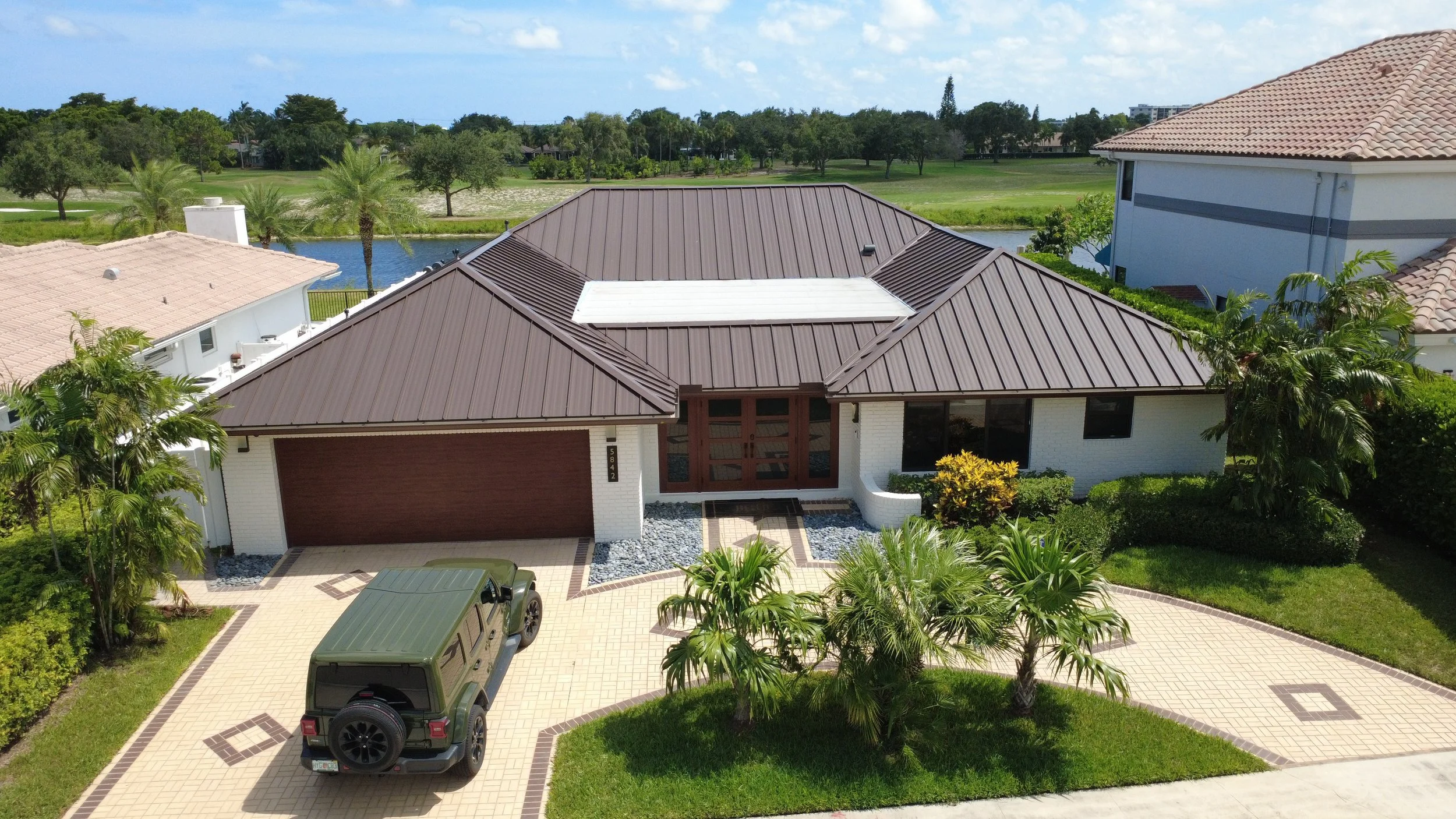 Aerial view of a modern house with a brown metal roof, surrounded by greenery, with a driveway and two vehicles parked outside. There is a pond and trees in the background.