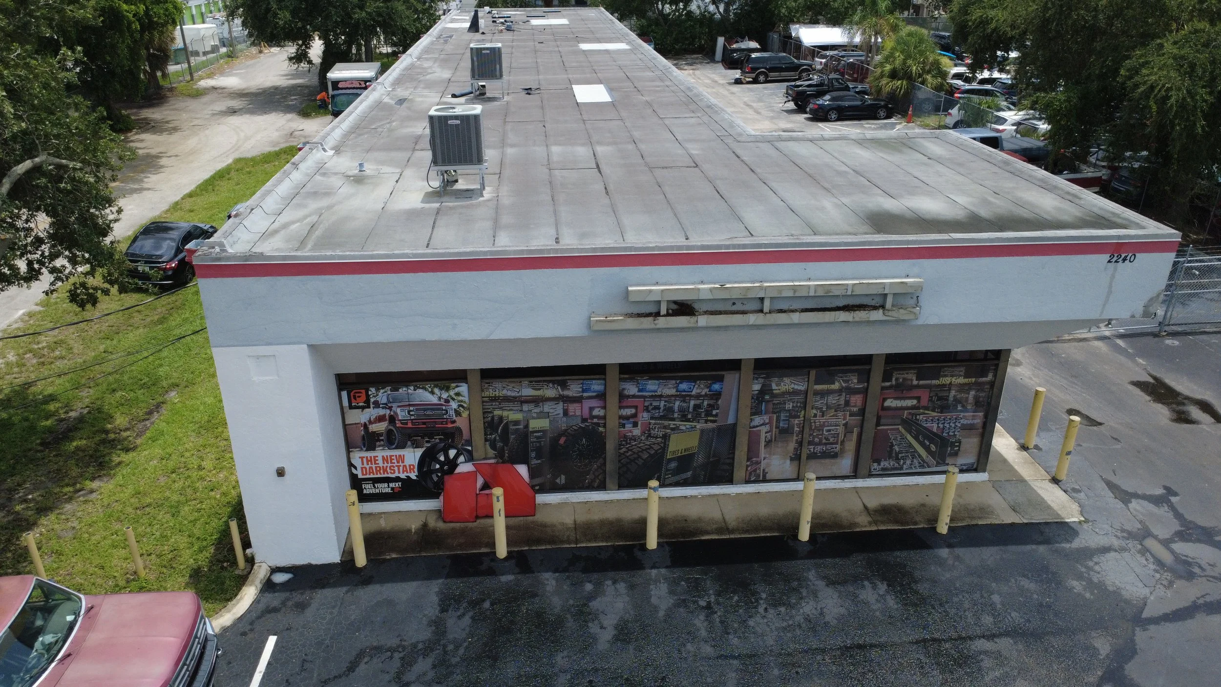 An aerial view of a commercial building with a flat roof, air conditioning units, parking lot, and cars parked around it.