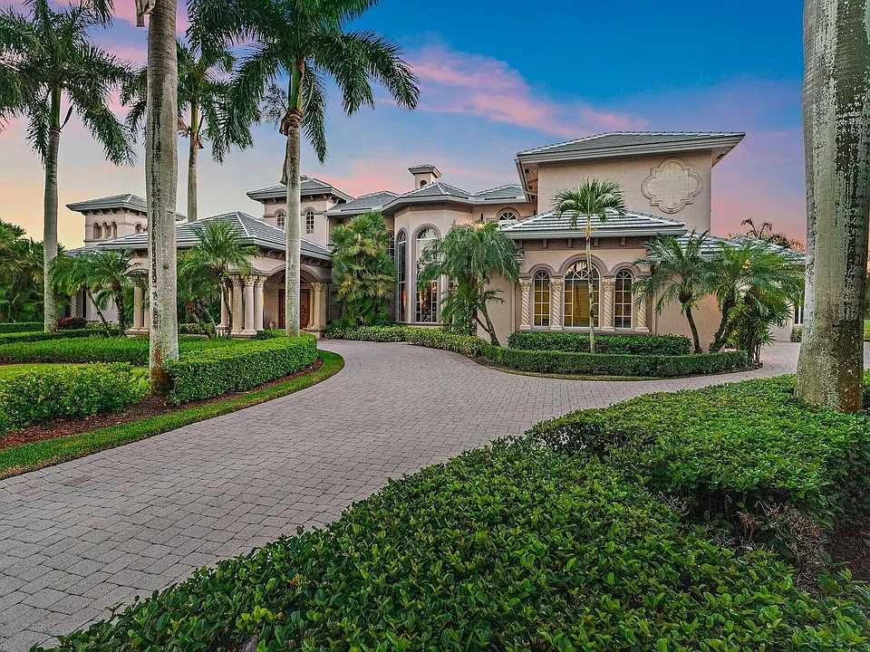 Large upscale mansion with beige walls, surrounded by palm trees and landscaped bushes, with a paved driveway leading up to the house, under a colorful sunset sky.