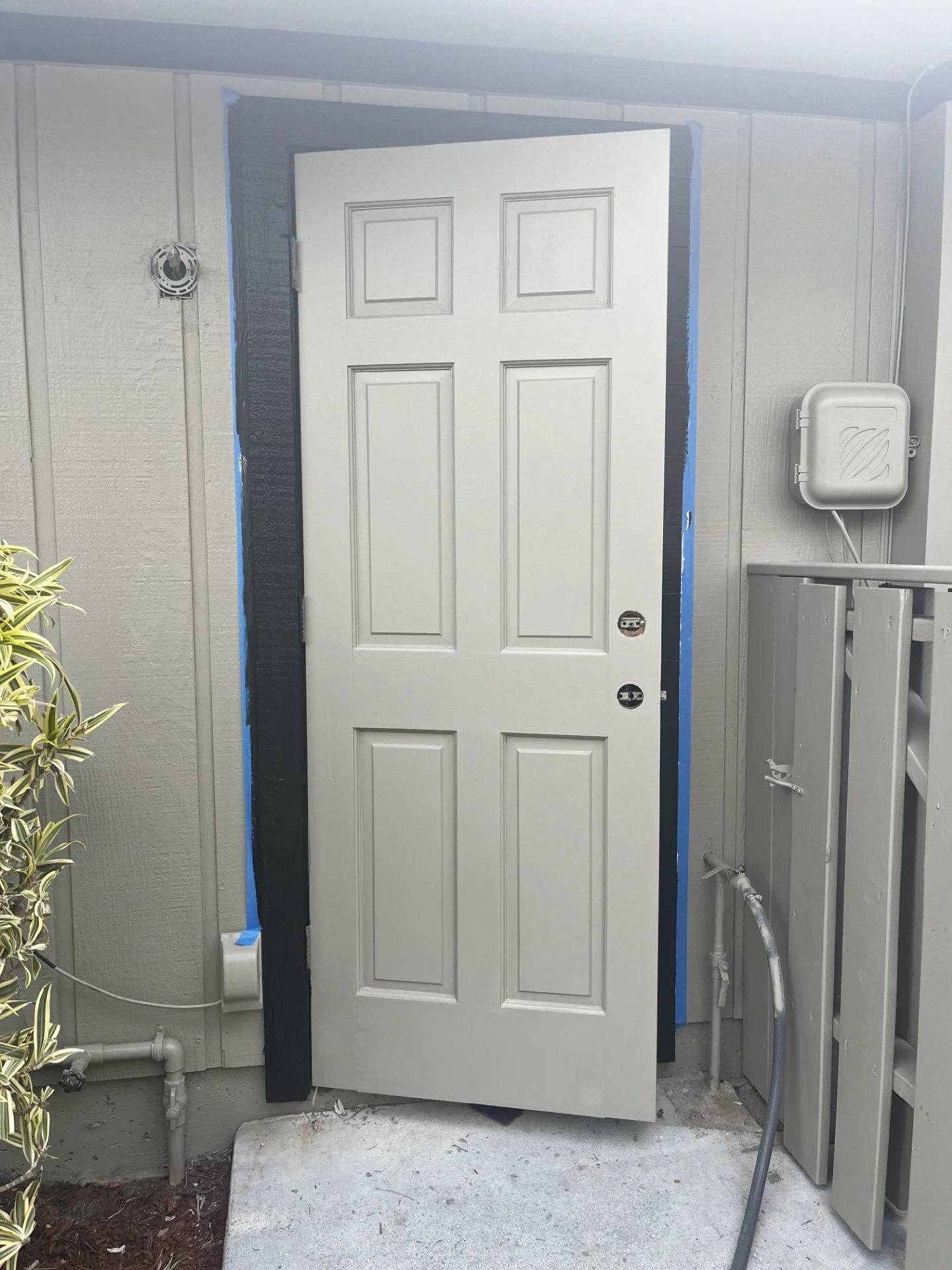 Unfinished white exterior door hanging off hinges on a house, with a blue painter's tape frame and an electrical box to the right, on a concrete porch with plants on the left.