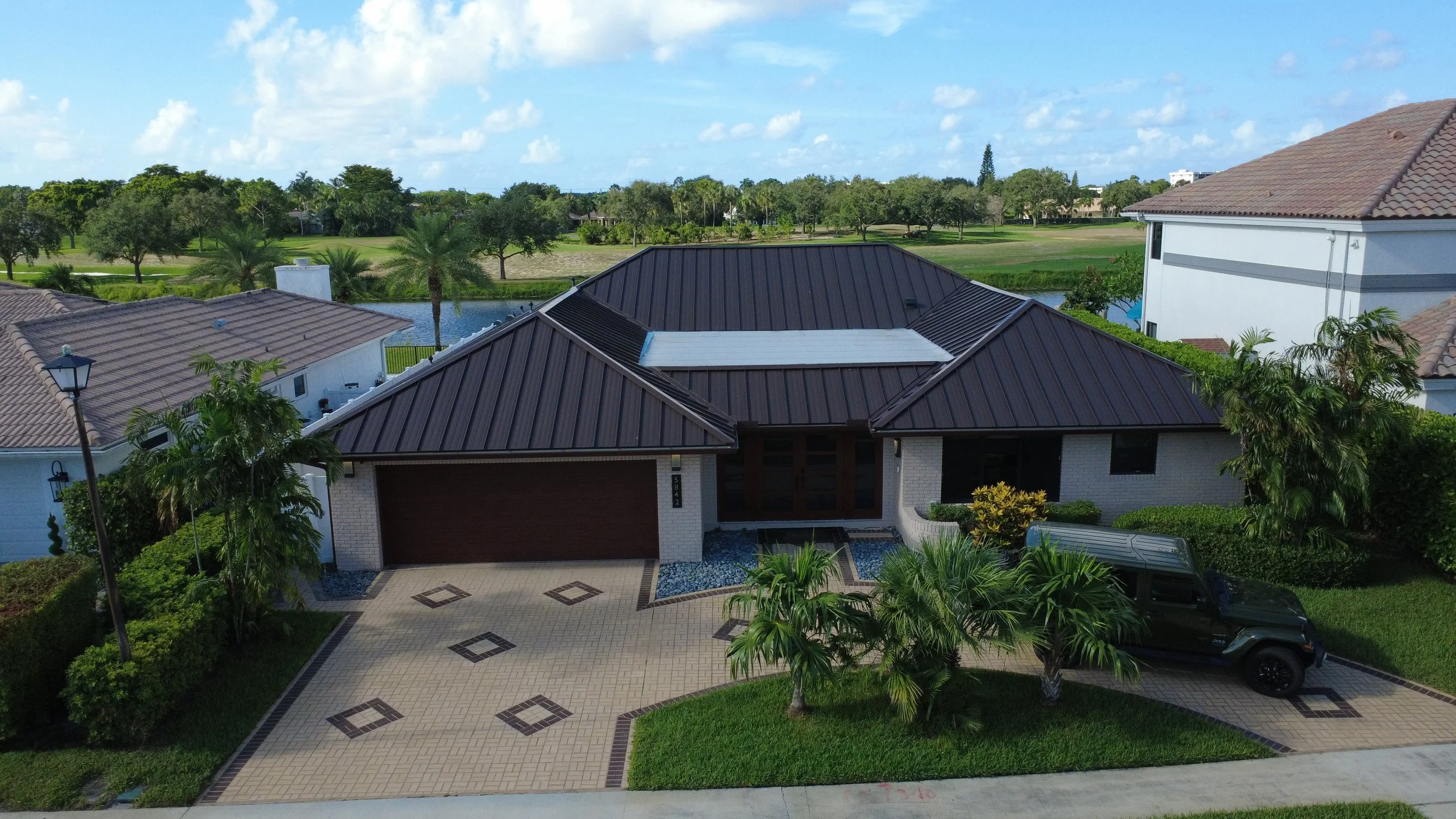 A modern white house with a dark metal roof, front driveway with decorative pattern, palm trees, and lush green landscaping in a suburban neighborhood with water and trees in the background under cloudy blue sky.