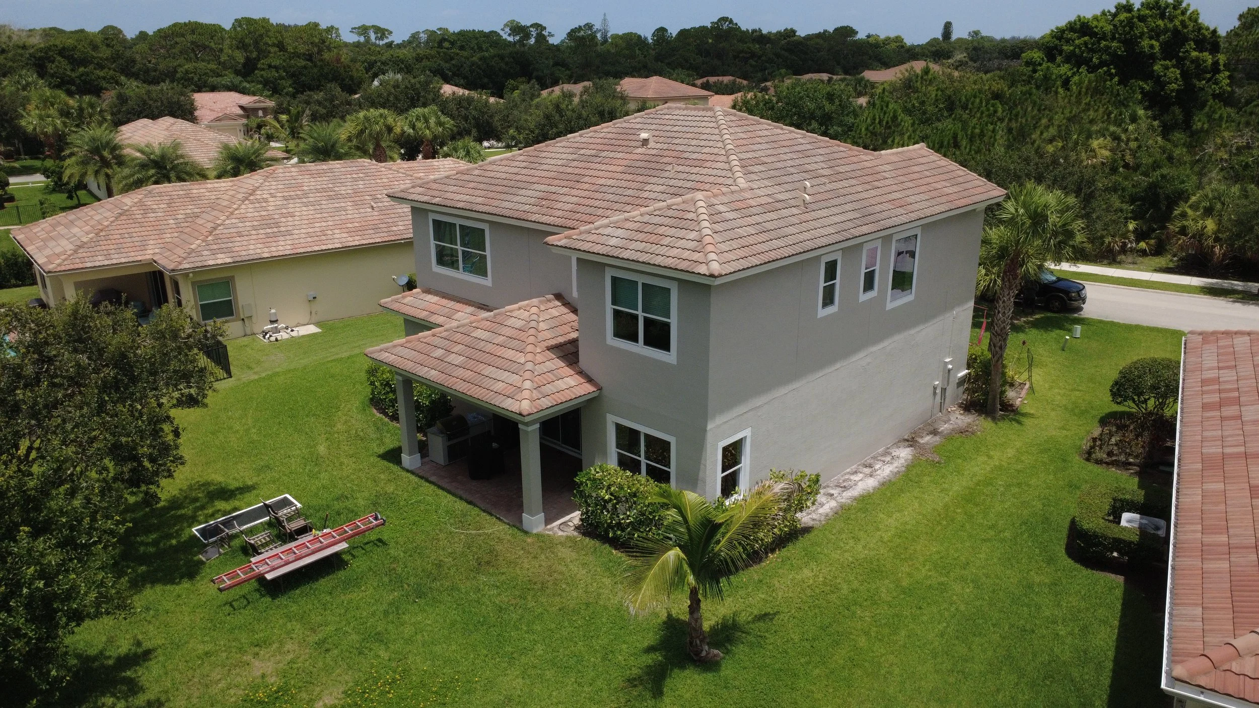 An aerial view of a two-story house with a red tile roof, surrounded by lush green yard and nearby trees in a neighborhood.