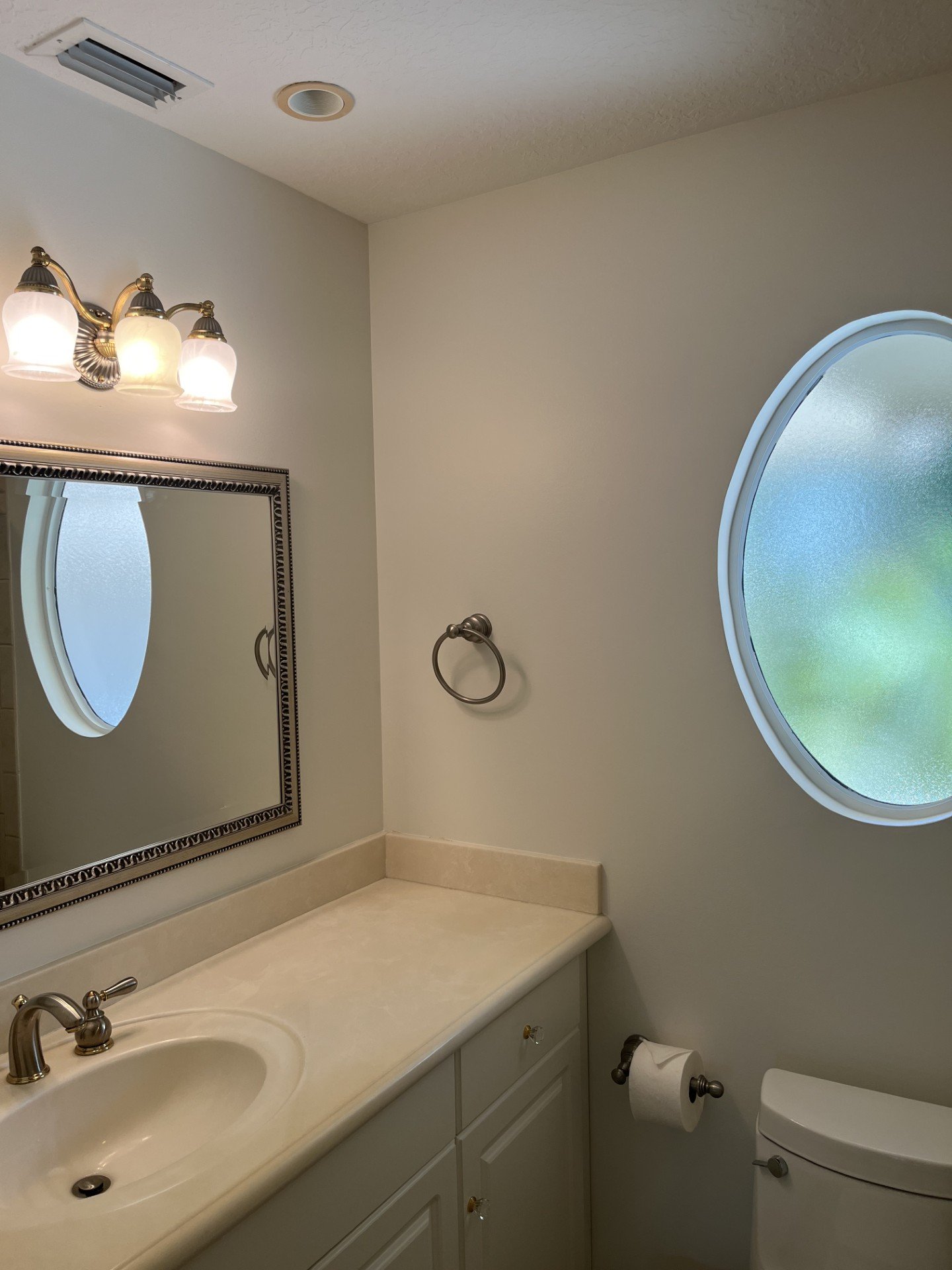 A bathroom with a white countertop, oval mirror, three-light vanity fixture, frosted oval window, towel ring, and toilet paper holder.