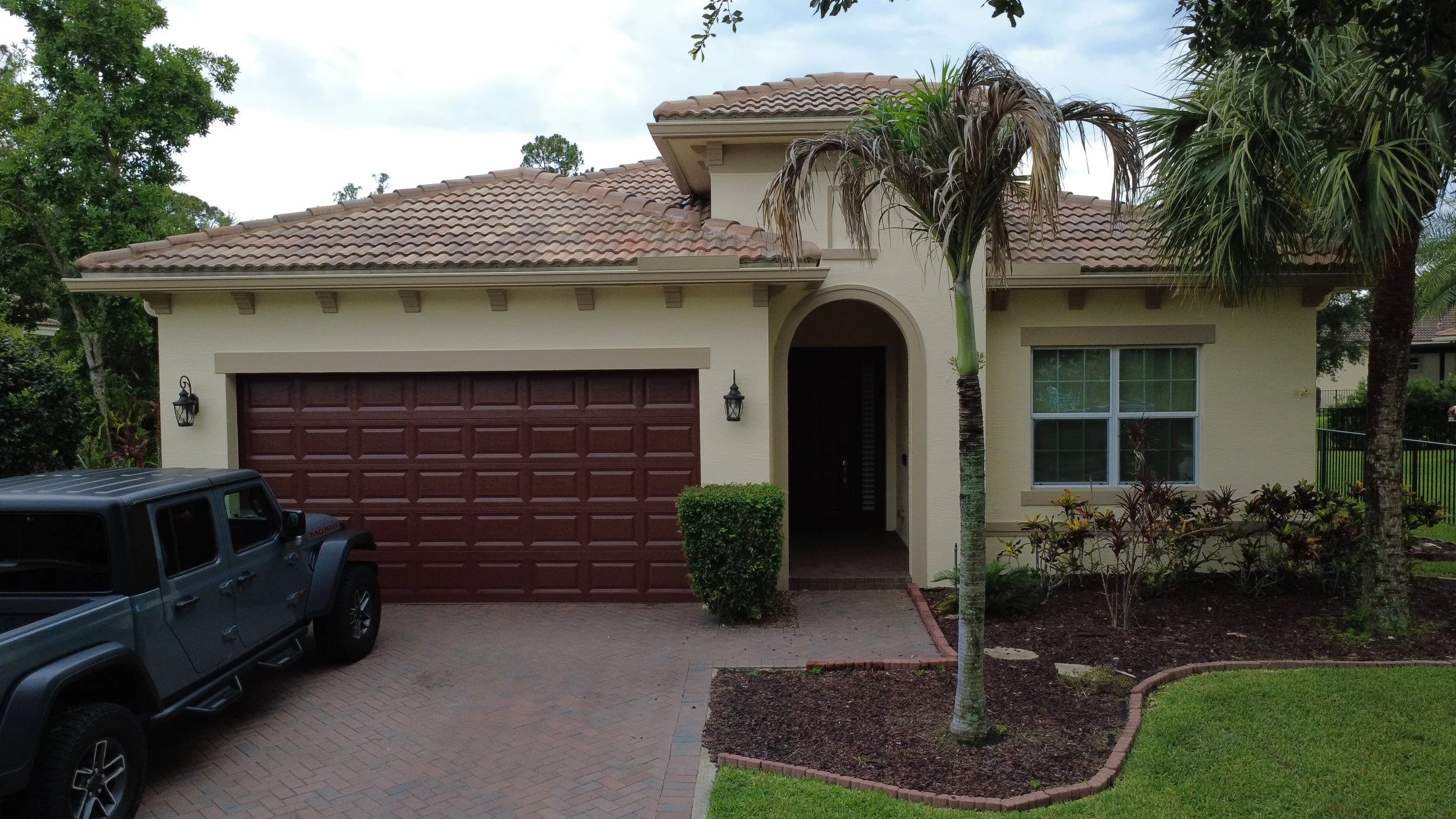 Front view of a beige house with a red garage door, a palm tree in the front yard, and a Jeep parked on the driveway.