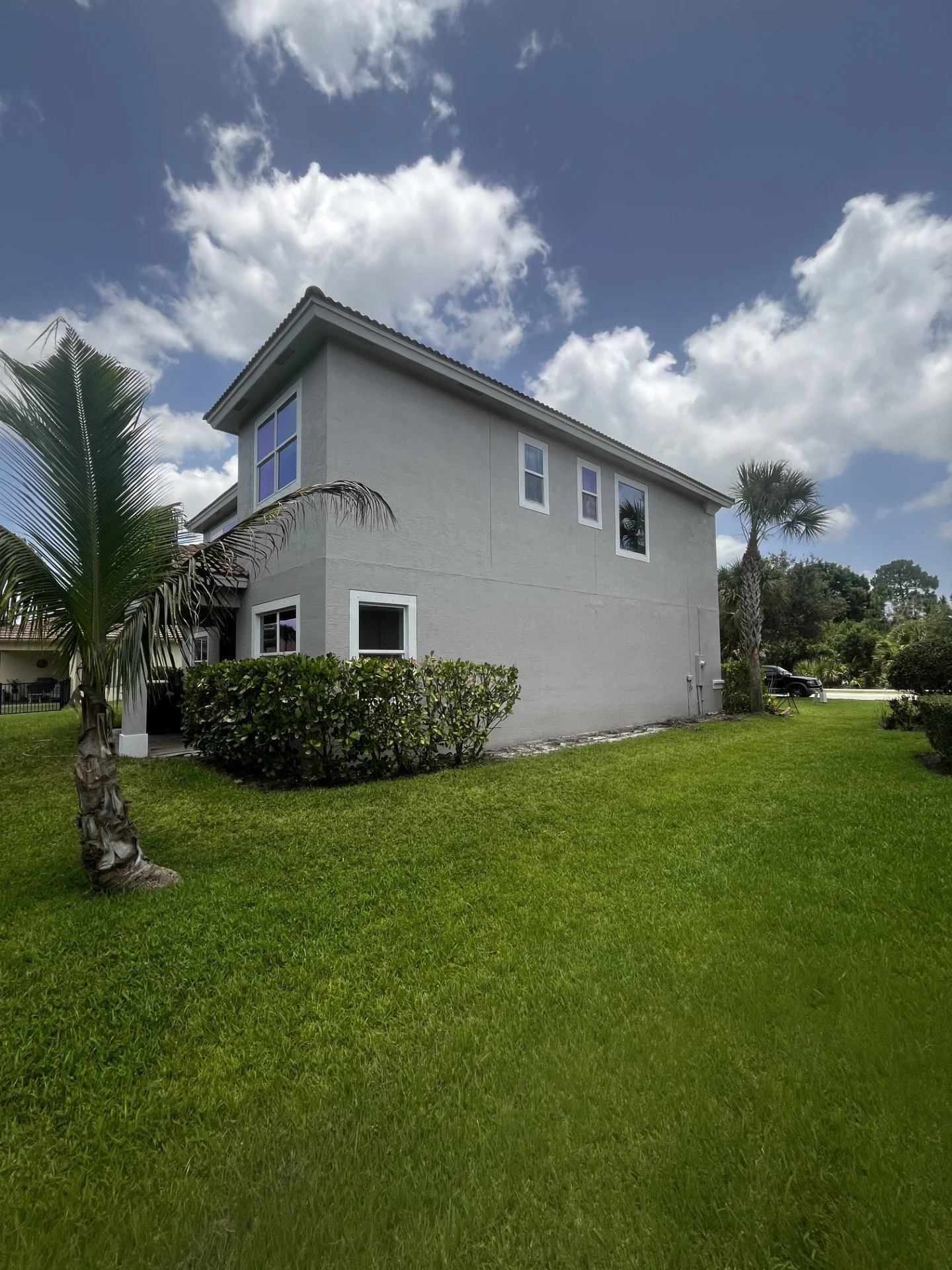 A two-story white house with multiple windows, surrounded by green lawn, palm trees, and bushes, under a partly cloudy sky.