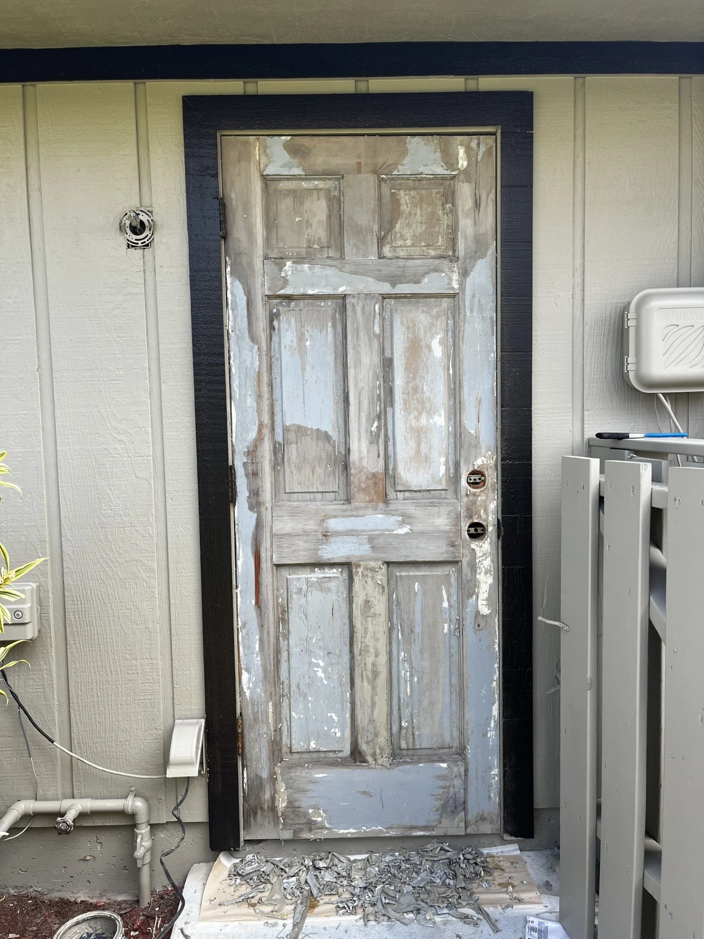 Old wooden door with peeling paint, surrounded by beige and black siding, with debris and broken paint chips on the ground below.