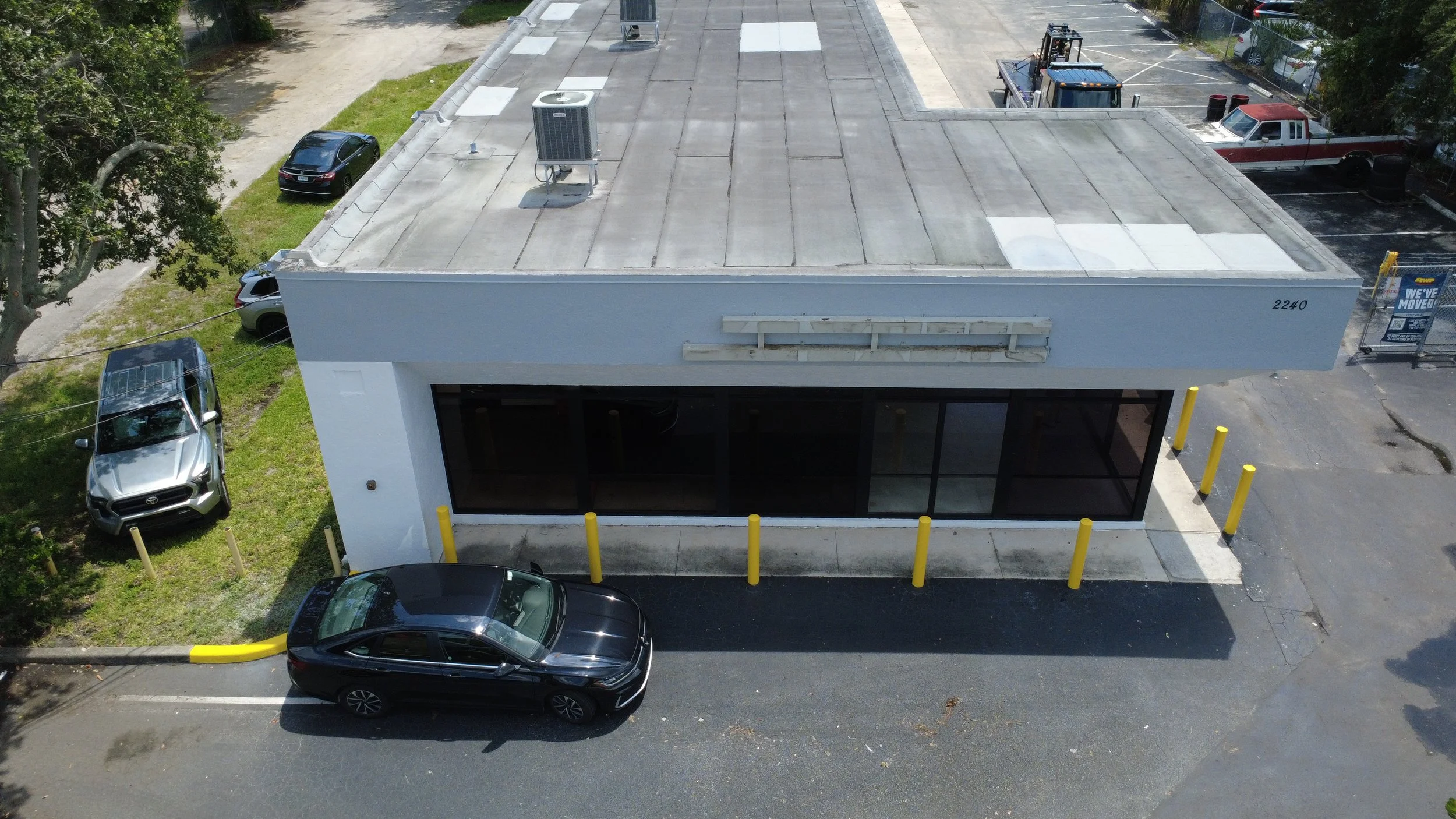 An aerial view of a white commercial building with a flat roof, air conditioning units, and large windows, surrounded by parking for cars and trucks, with some yellow bollards at the entrance.