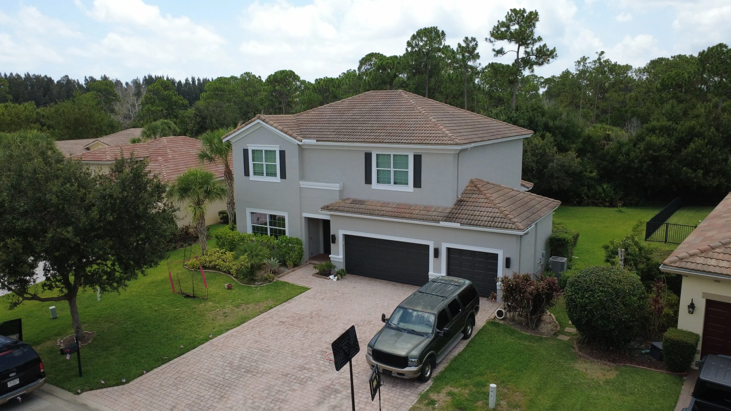 A two-story house with a gray exterior and a tiled roof, located in a green suburban neighborhood with trees, a lawn, and parked cars in the driveway.