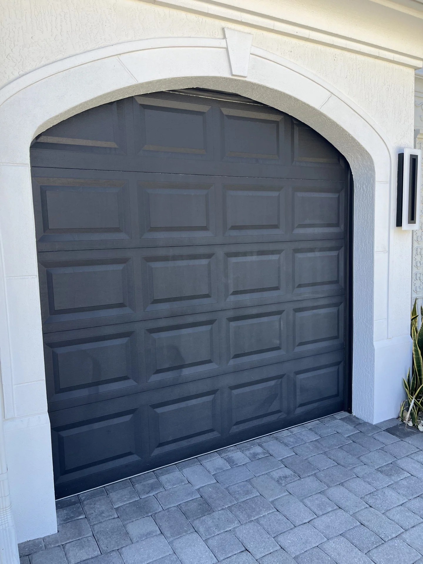 A closed dark gray garage door with rectangular panel design, surrounded by a white textured stucco wall and a white arched frame, with a gray brick driveway in front.