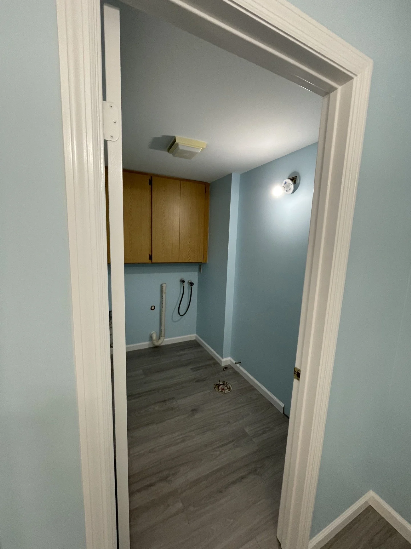 Empty laundry room with blue walls, wooden cabinets, and plumbing hookups for a washer and dryer, with gray wood-look flooring.