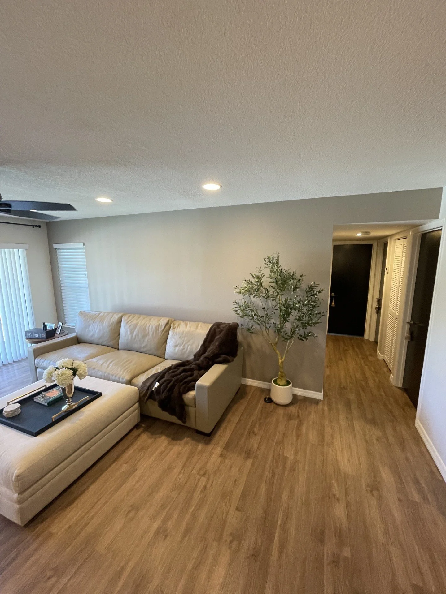Living room with beige sectional sofa, coffee table with flower arrangement, potted plant, and beige walls. There are windows with blinds and a dark door in the background.