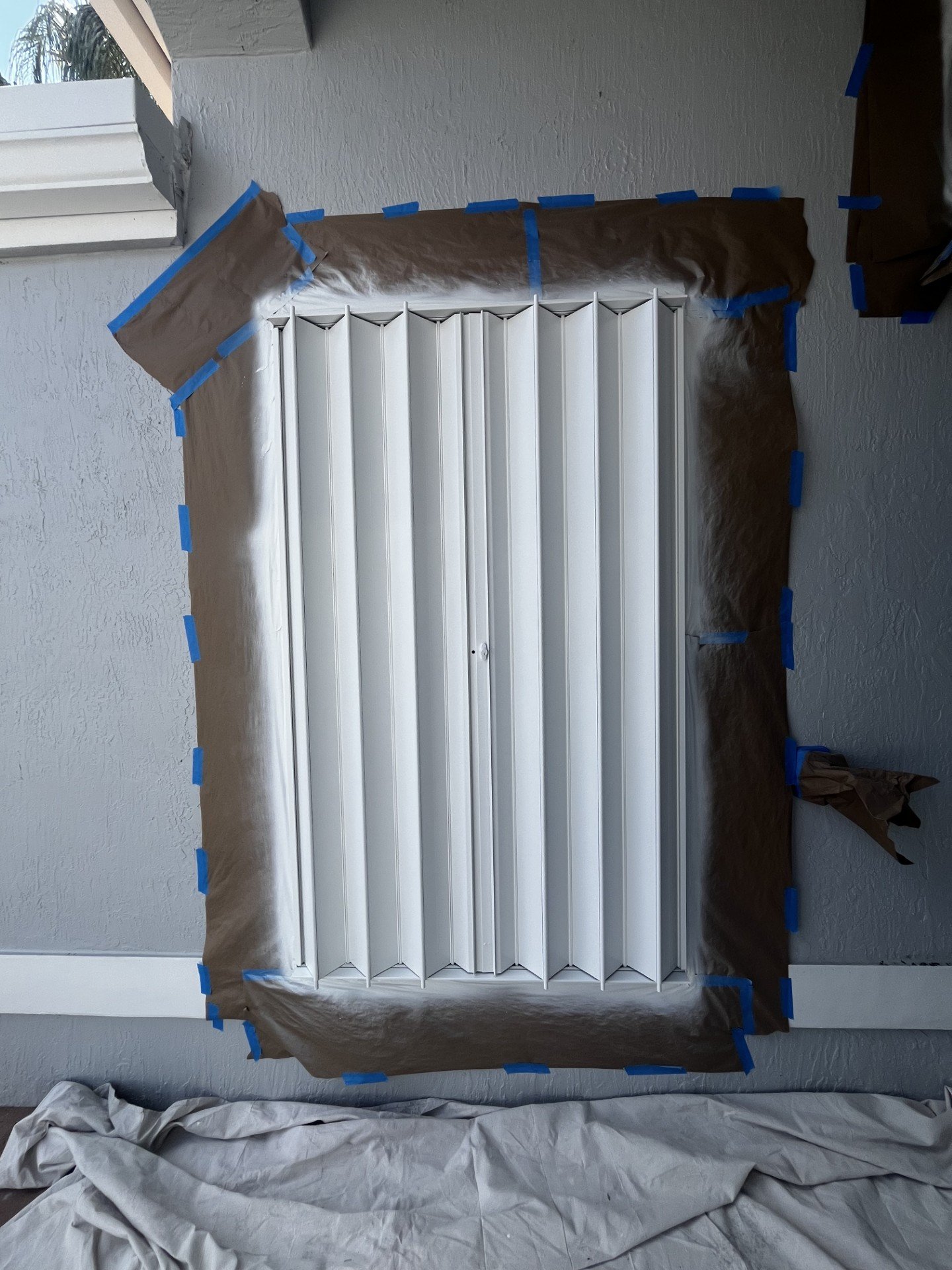 A wall-mounted white folding closet door framed with brown painter's paper and blue painter's tape, indicating preparation for painting.