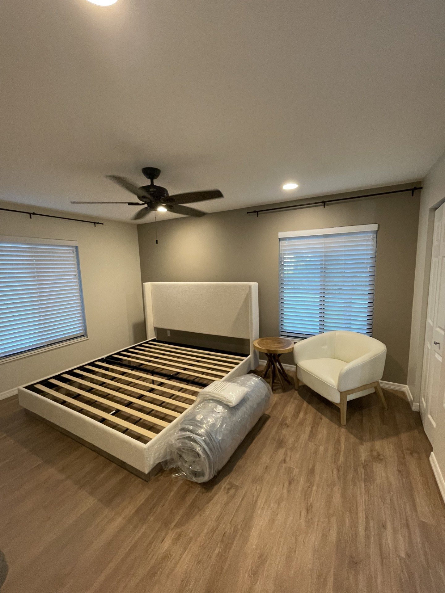 A bedroom with a bed frame but no mattress, two windows with blinds, a ceiling fan, a white armchair, a small wooden table, and a rolled-up plastic mattress cover.