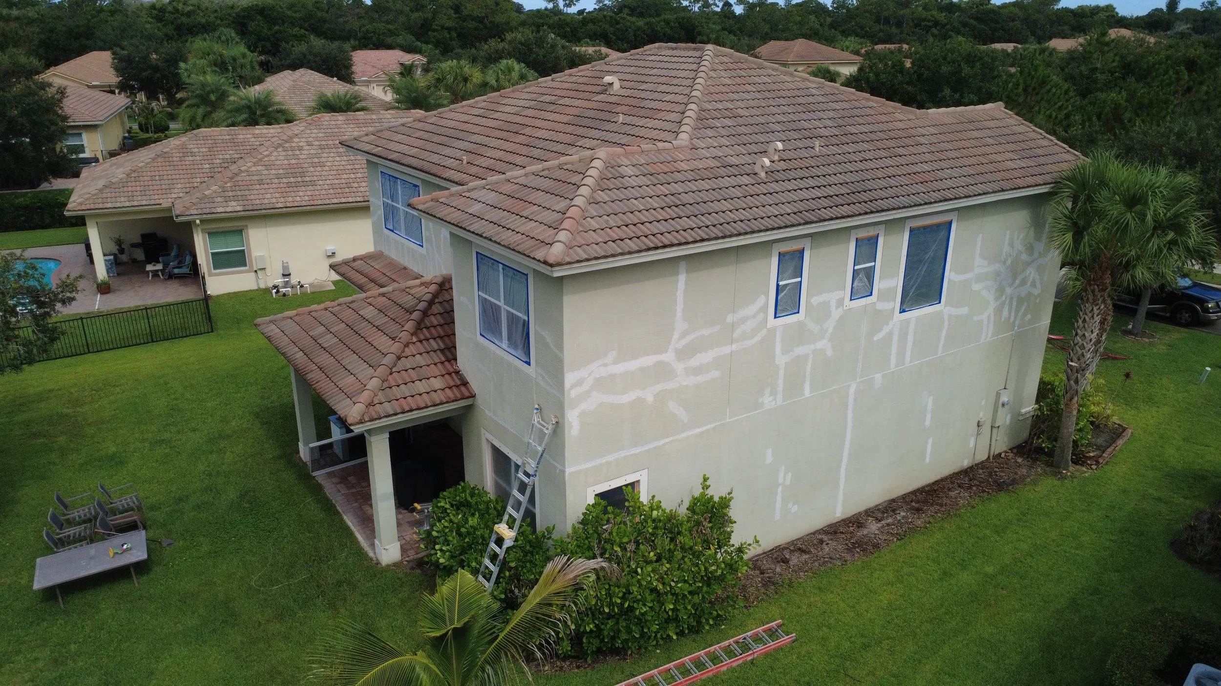 A two-story house under construction with a tiled roof, partially finished exterior walls, and a ladder leaning against the side. The backyard features a green lawn, outdoor furniture, a small pool area, and surrounding trees.