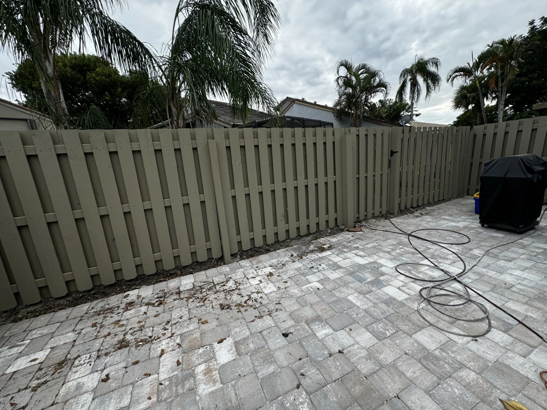 Outdoor backyard patio with a beige wooden fence, tropical palm trees, cloudy sky, and black covered grill on stone paver surface.