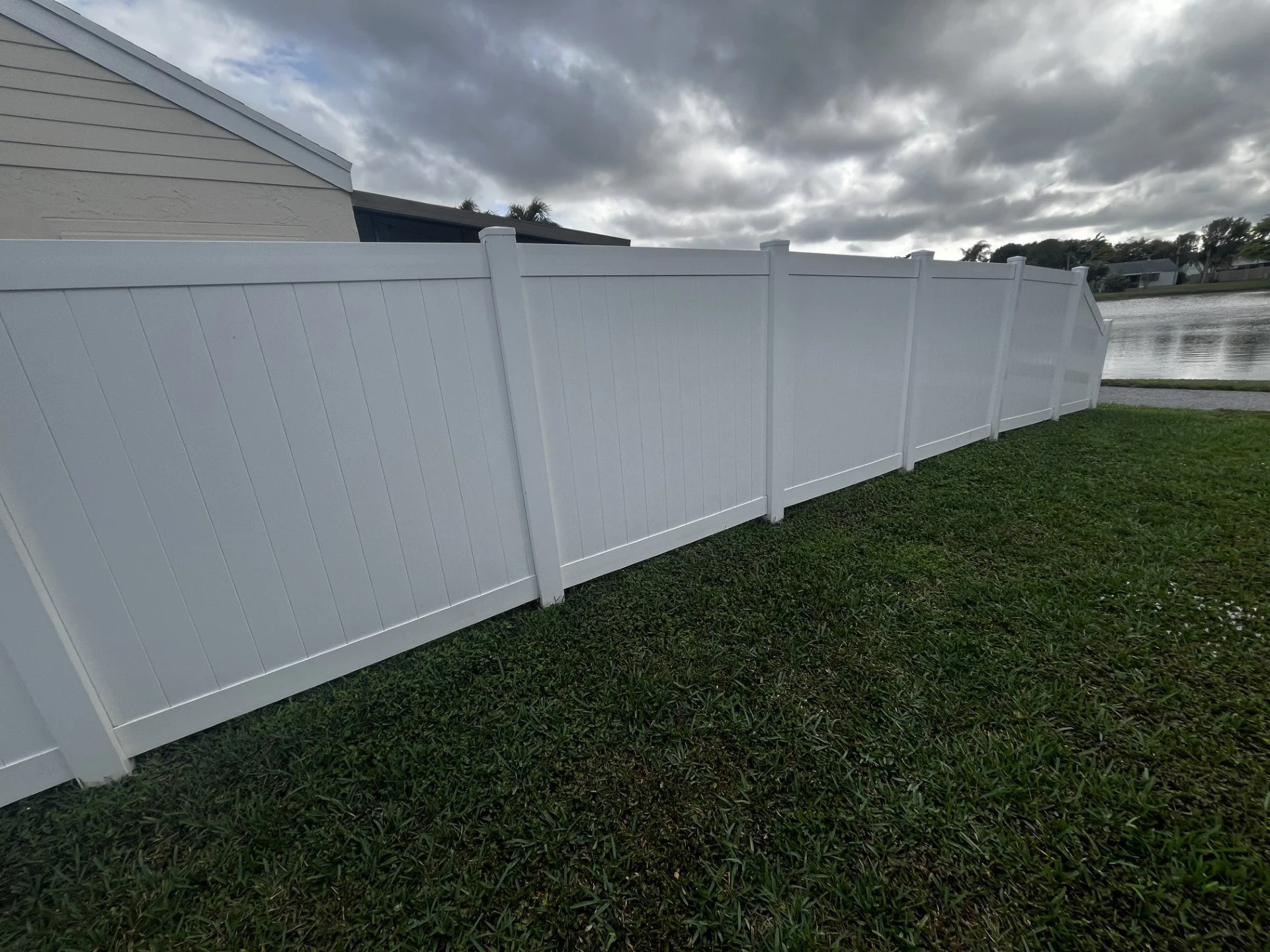 A white vinyl privacy fence running alongside a house by a body of water, under cloudy skies with dark clouds overhead.