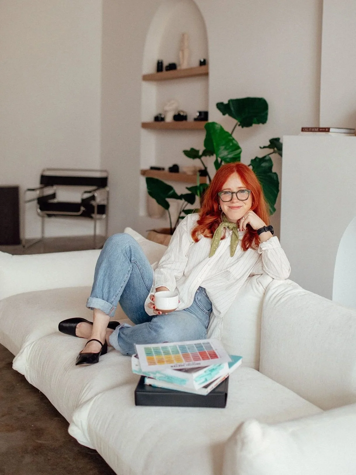 Haleigh Culverhouse sitting on a cream-colored sofa, holding a white cup, smiling, wearing a striped white shirt, blue jeans, and black shoes with ankle straps in a modern living room with green plants, shelves, and a black chair in the background.