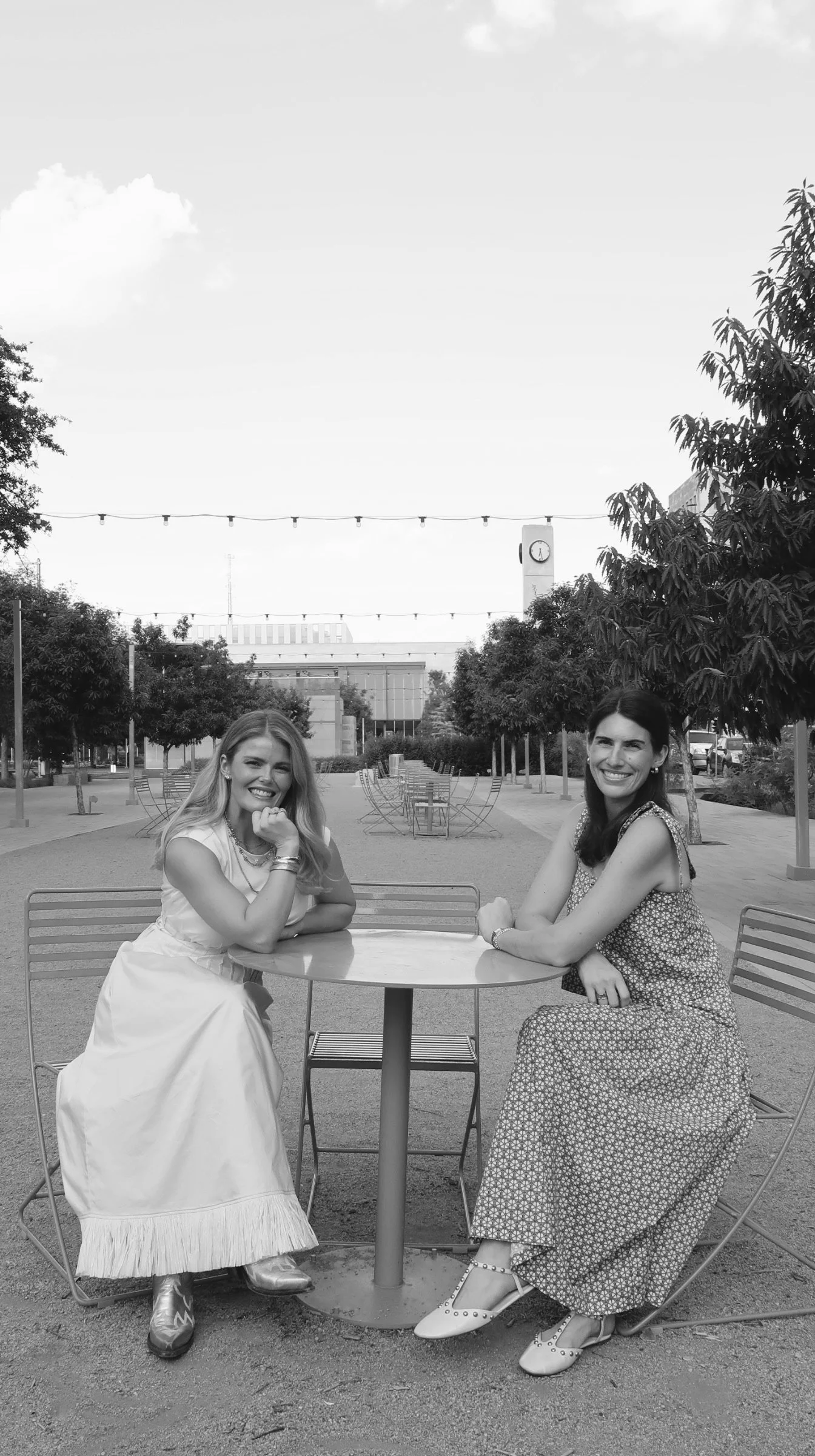 Two women sitting at a round outdoor table in a park, smiling at the camera, with trees and a clock tower in the background.